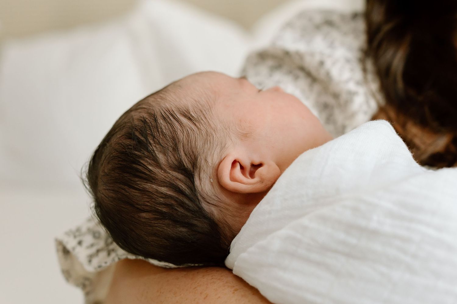 A sleeping newborn baby rests on white bedding with dark hair visible against the soft fabric.