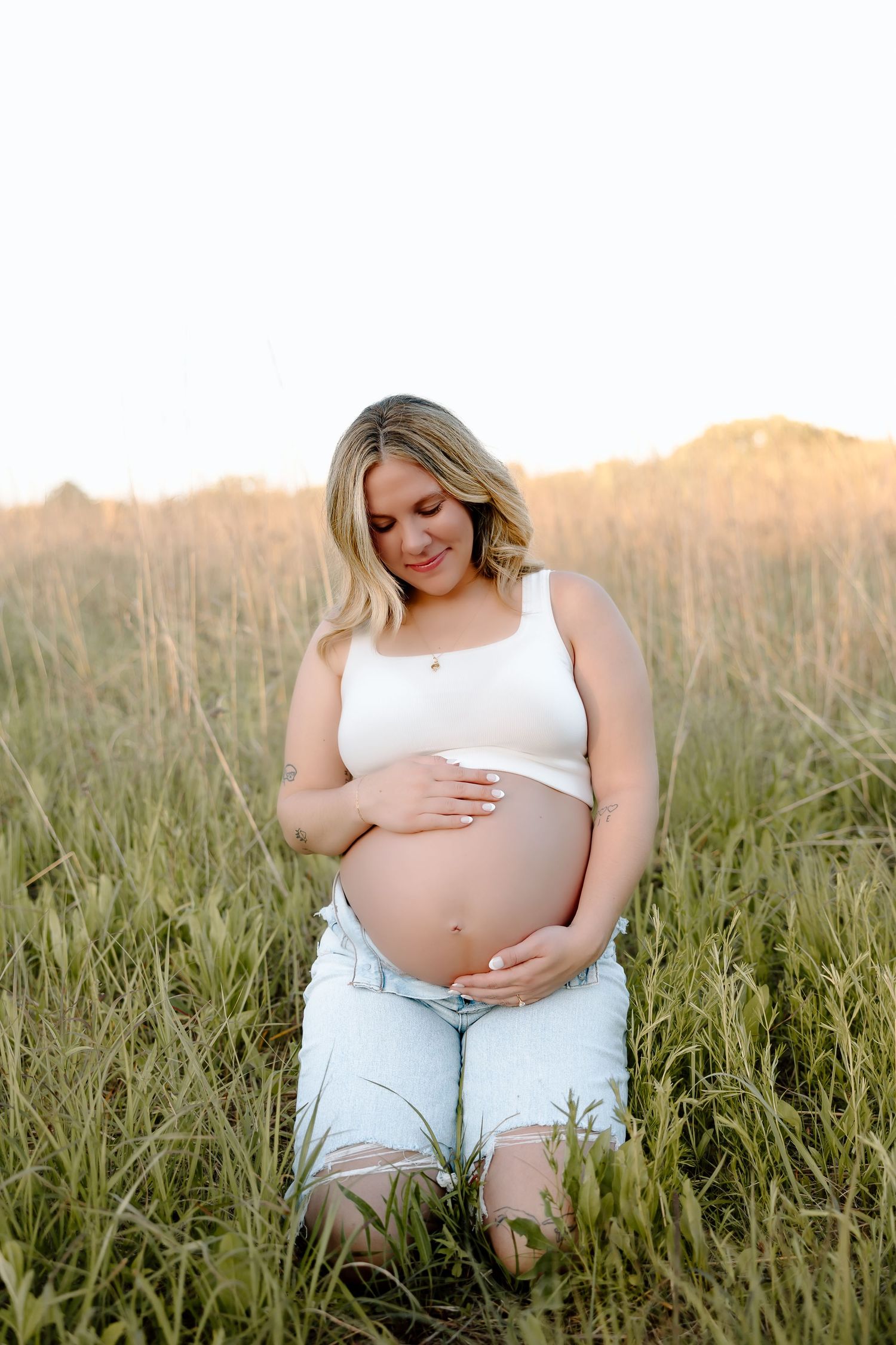 Maternity photos showing a pregnant woman in white sitting among tall grass at golden hour.