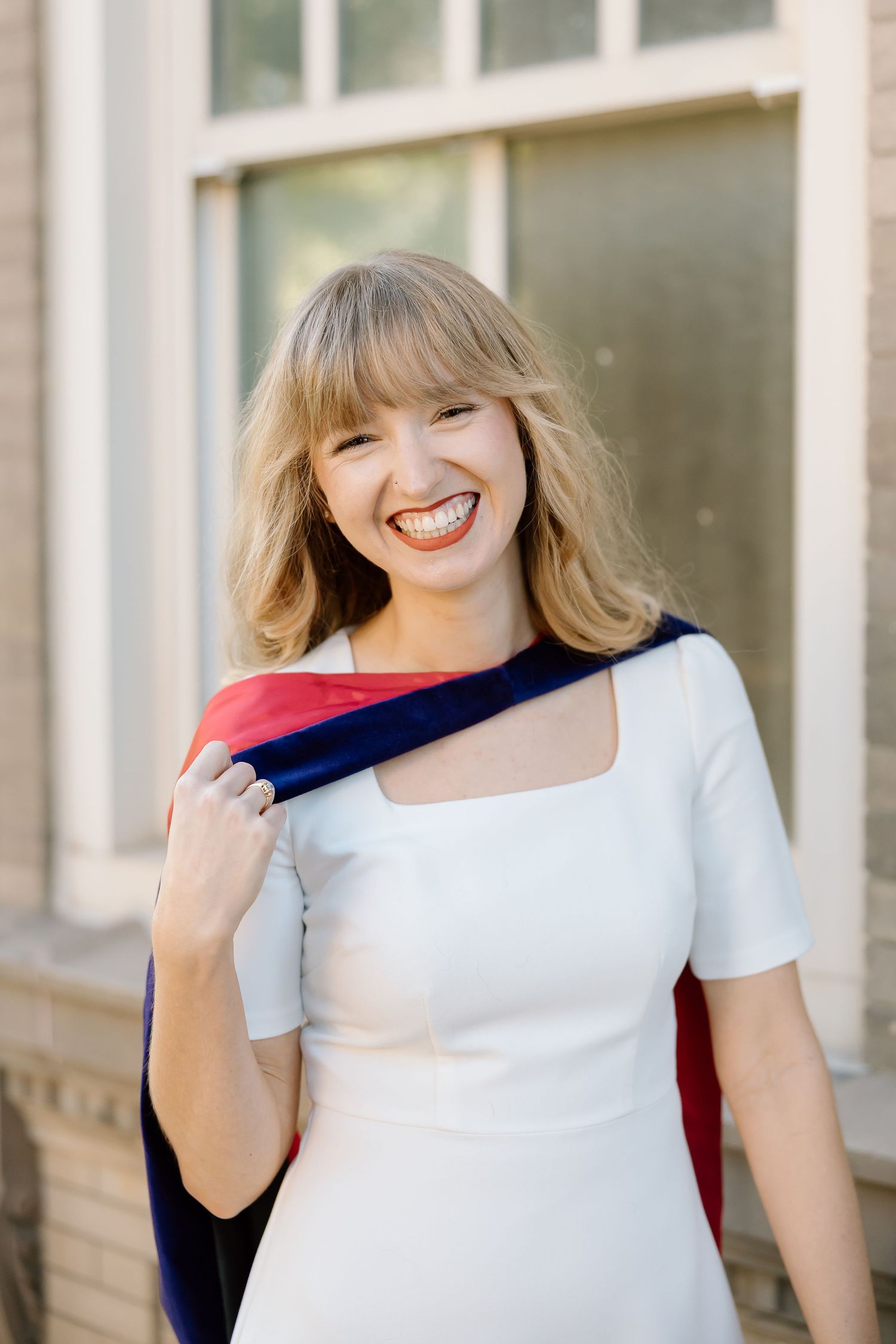 A person wearing a white top and dark cardigan stands against a light-colored wall with a bright smile.