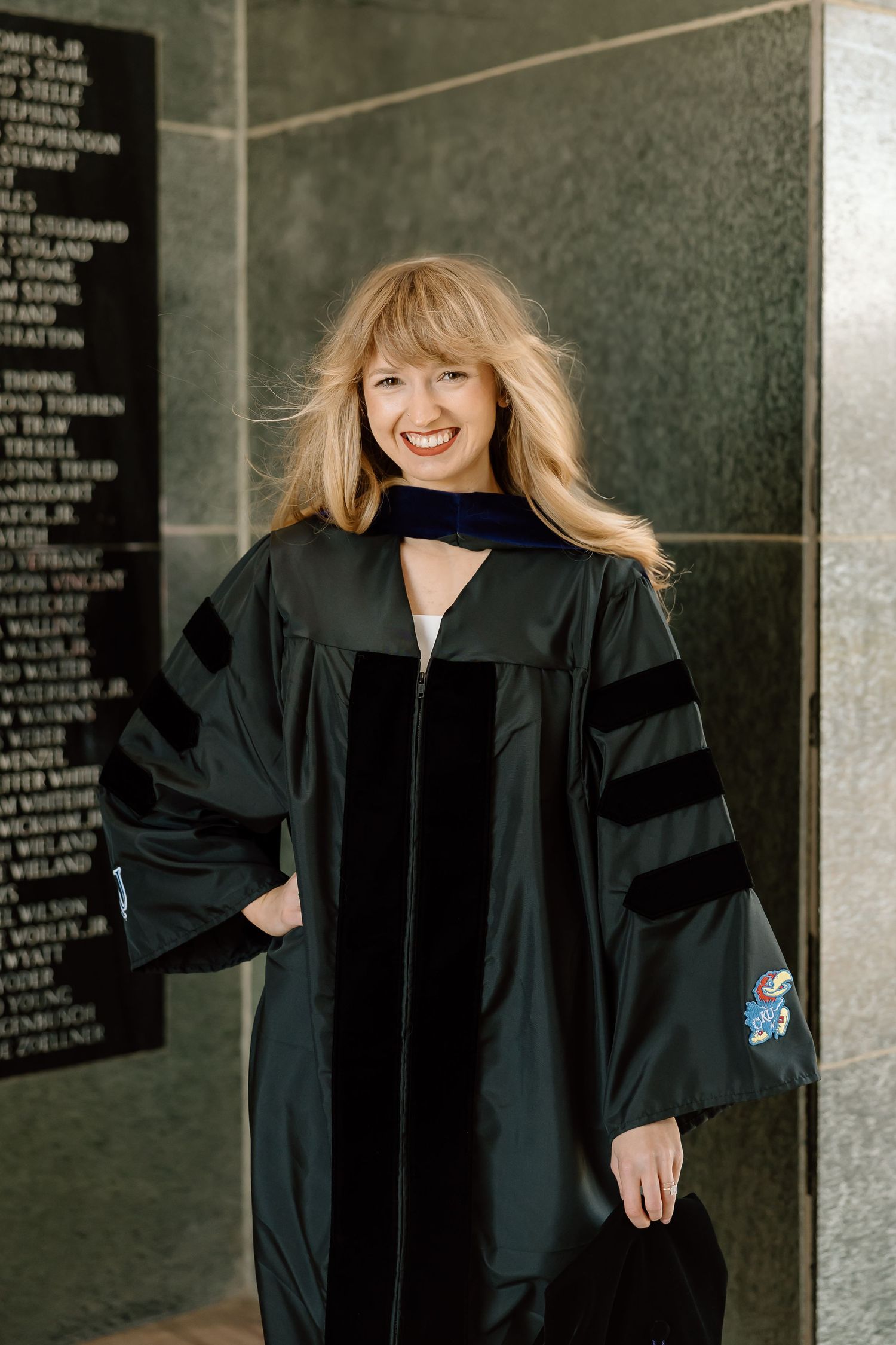 A graduate in a black academic gown poses joyfully against a wall with engraved text in three similar photos.