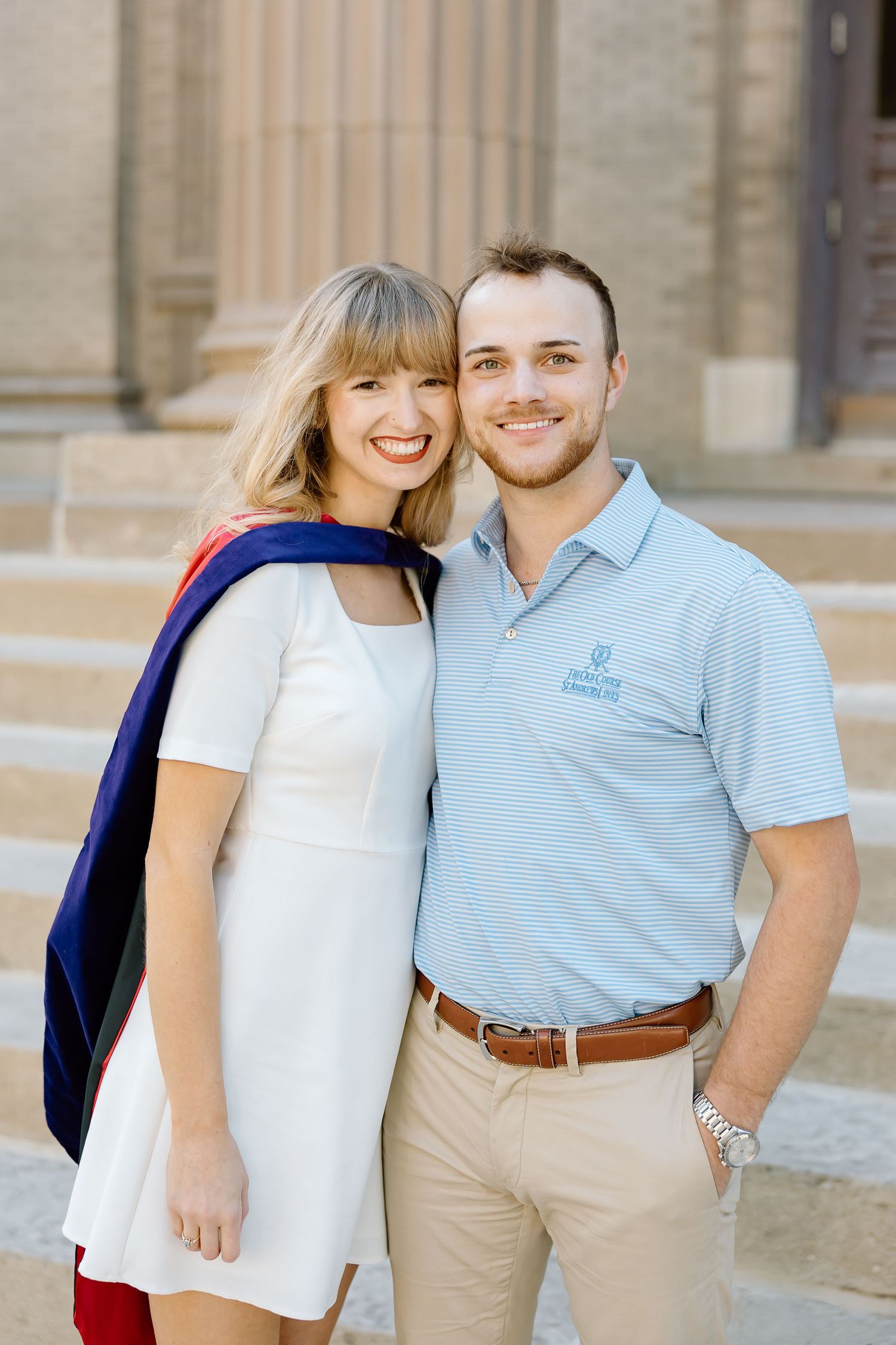 A couple in coordinating white and light blue polo shirts pose together against a stone building exterior.