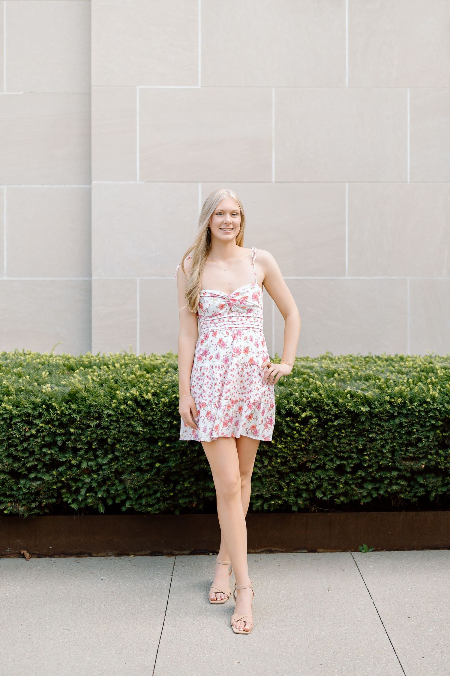 A person in a floral dress poses against a stone wall with manicured hedges.