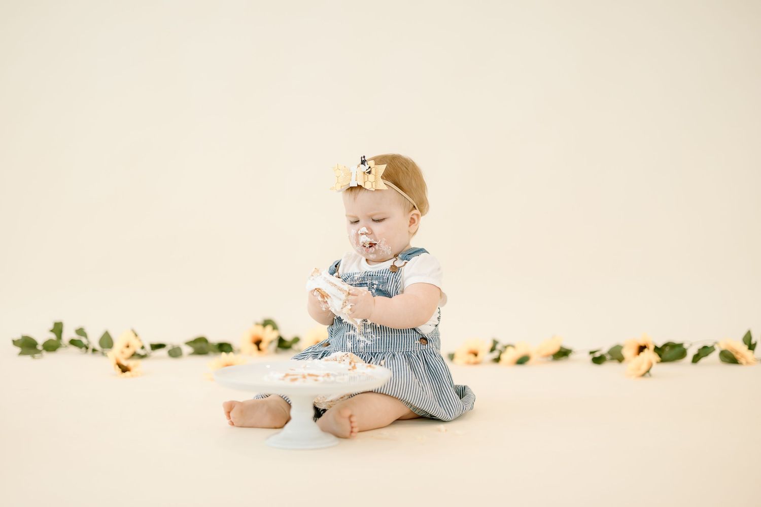 Baby wearing blue and white dress sits on cream backdrop with floral garland decoration.