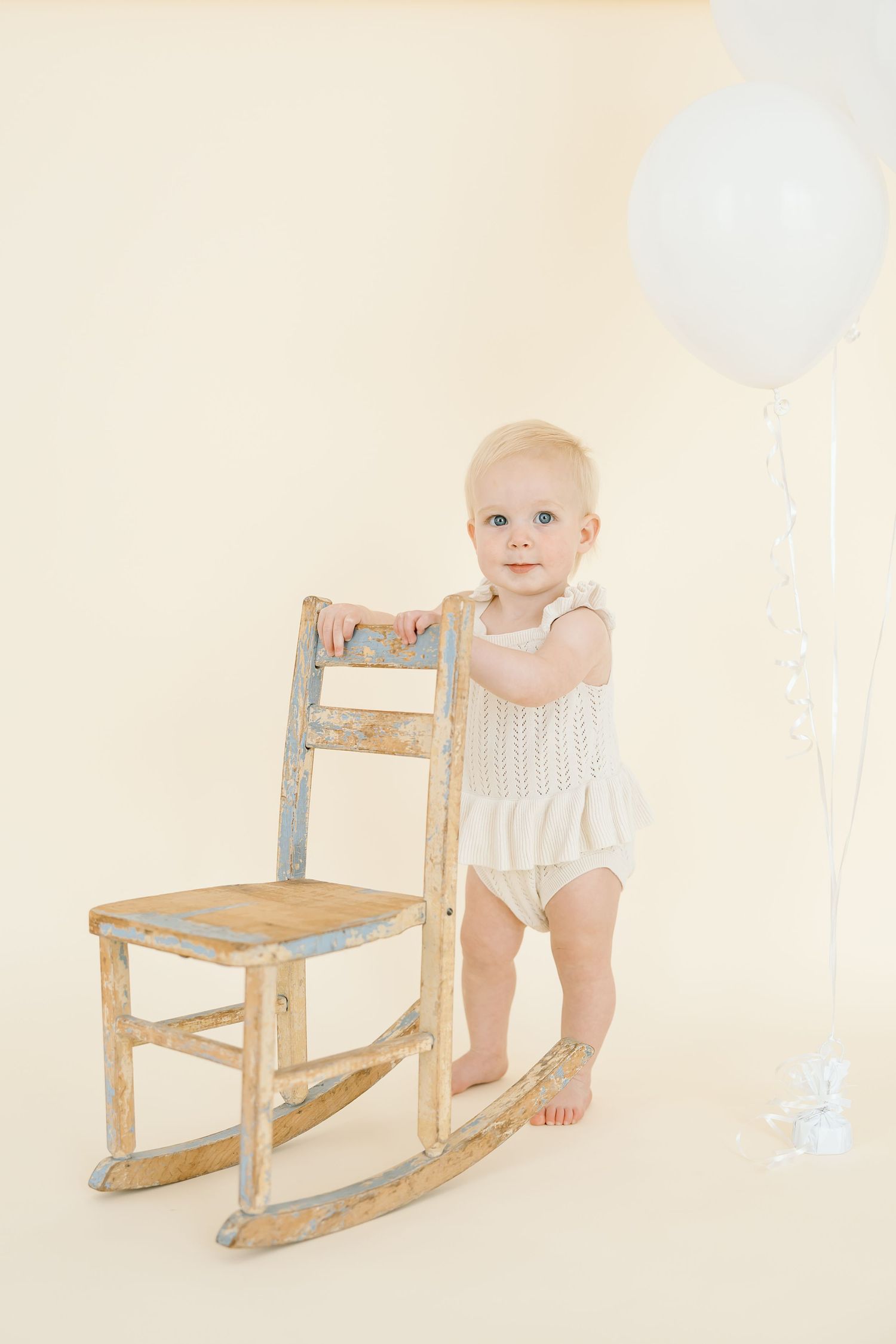 Blonde toddler in white romper standing next to a rustic wooden rocking chair with a white balloon nearby.