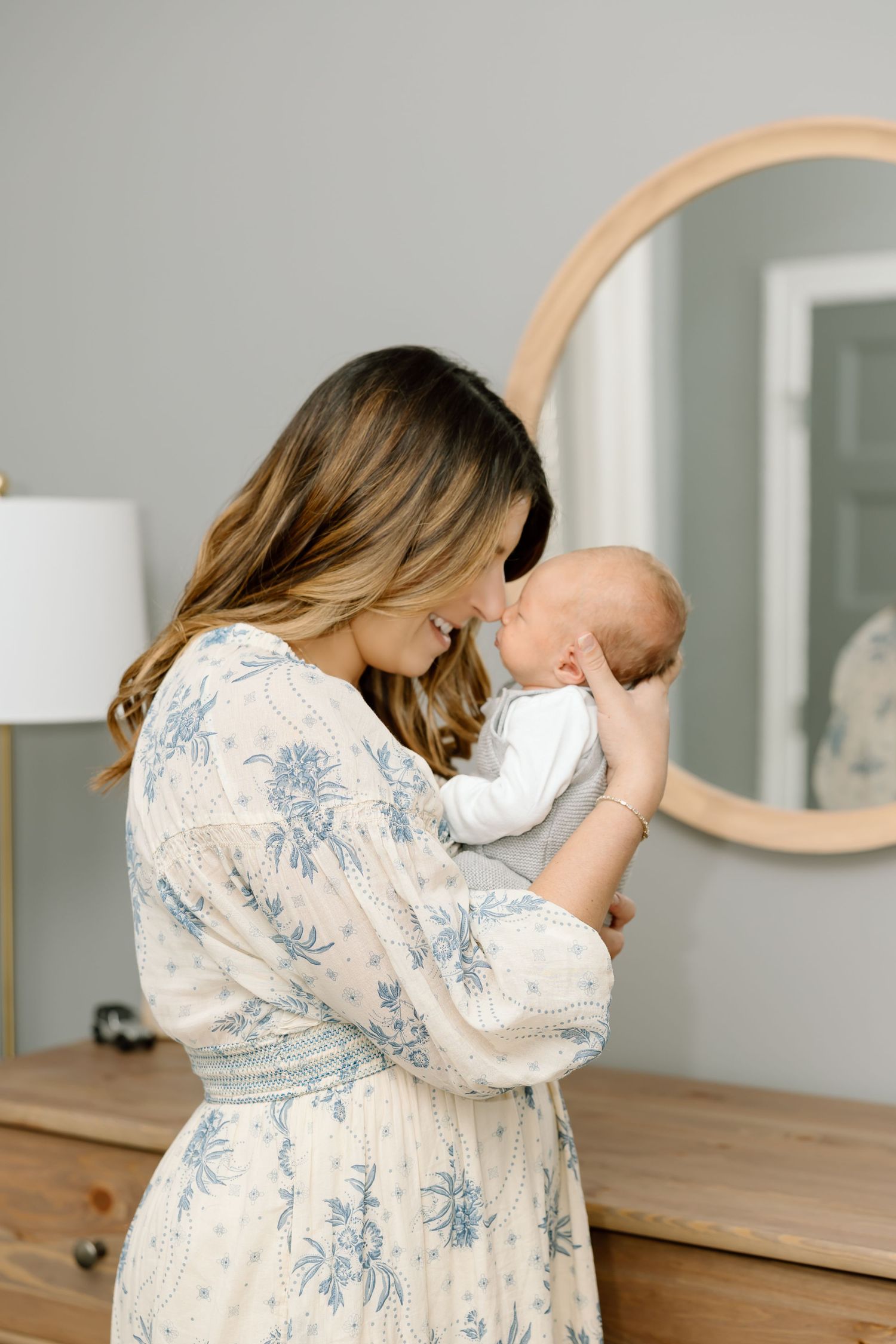 A tender moment captured as a mother in a floral dress cradles her newborn baby in a softly lit nursery with a round mirror.