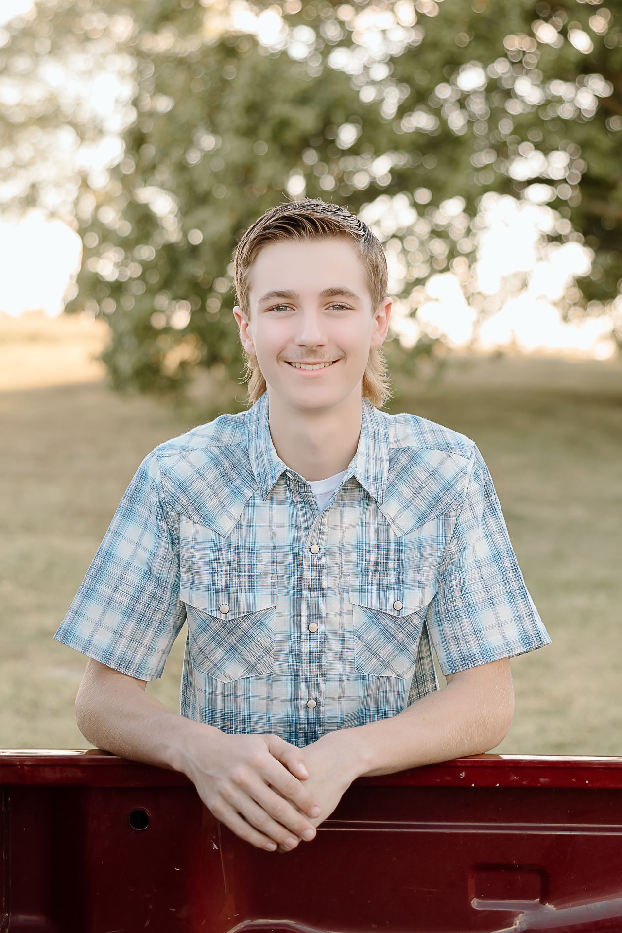 A person wearing a plaid blue and white short-sleeve western shirt leans against a red truck in an outdoor setting.