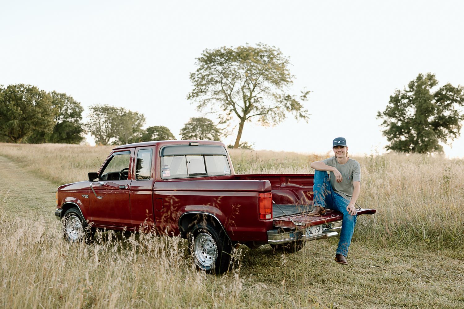 A red vintage pickup truck parked in a golden grass field with someone in blue clothing standing nearby.