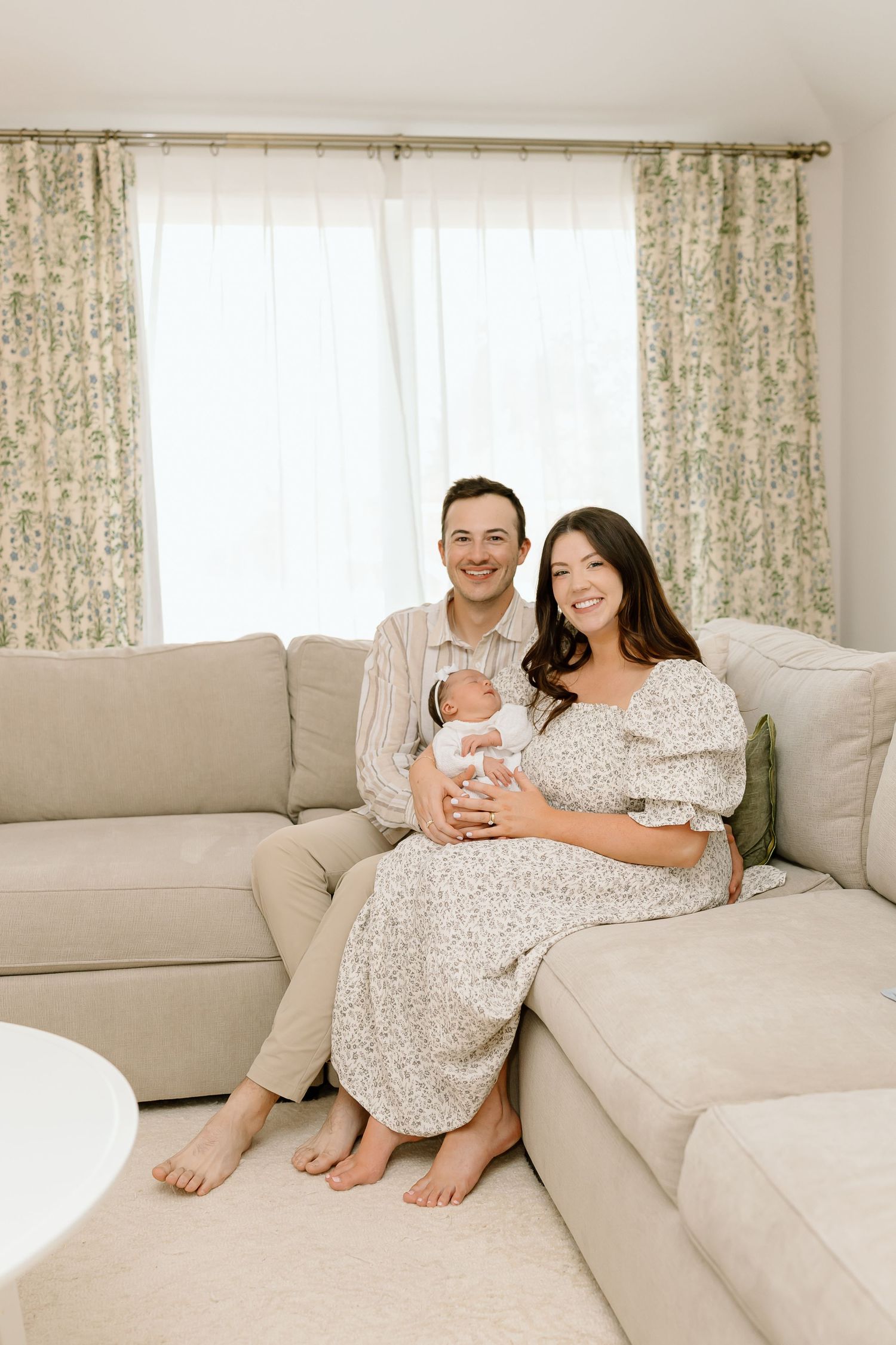 A family sits together on a beige sectional sofa in a bright living room with floral curtains.