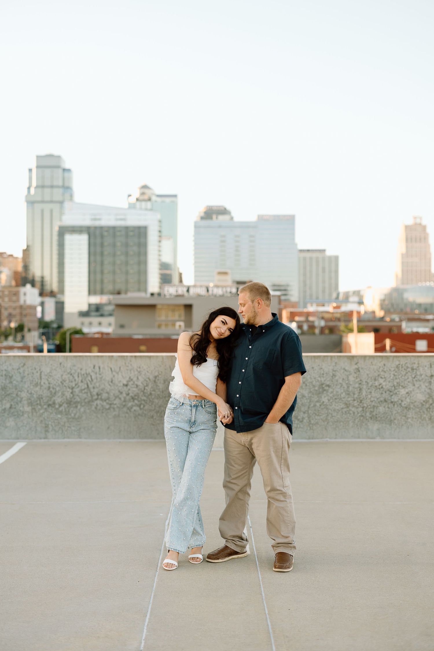 A couple stands together on a rooftop parking deck with the Kansas City skyline visible in the background at sunset.