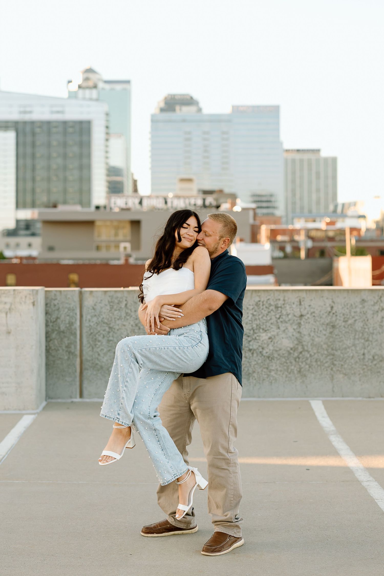A romantic couple embraces and dips on a rooftop parking lot with city buildings in the background at sunset.