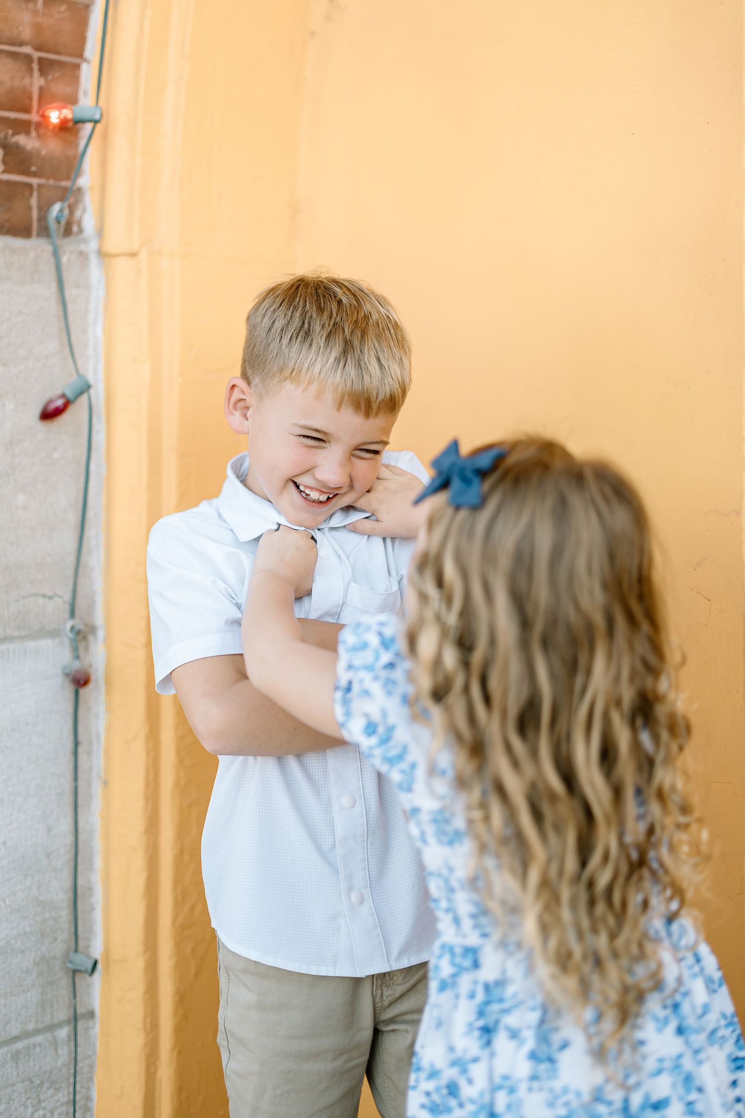 Two young children in a hallway share a playful moment as one covers their face while wearing a white shirt.
