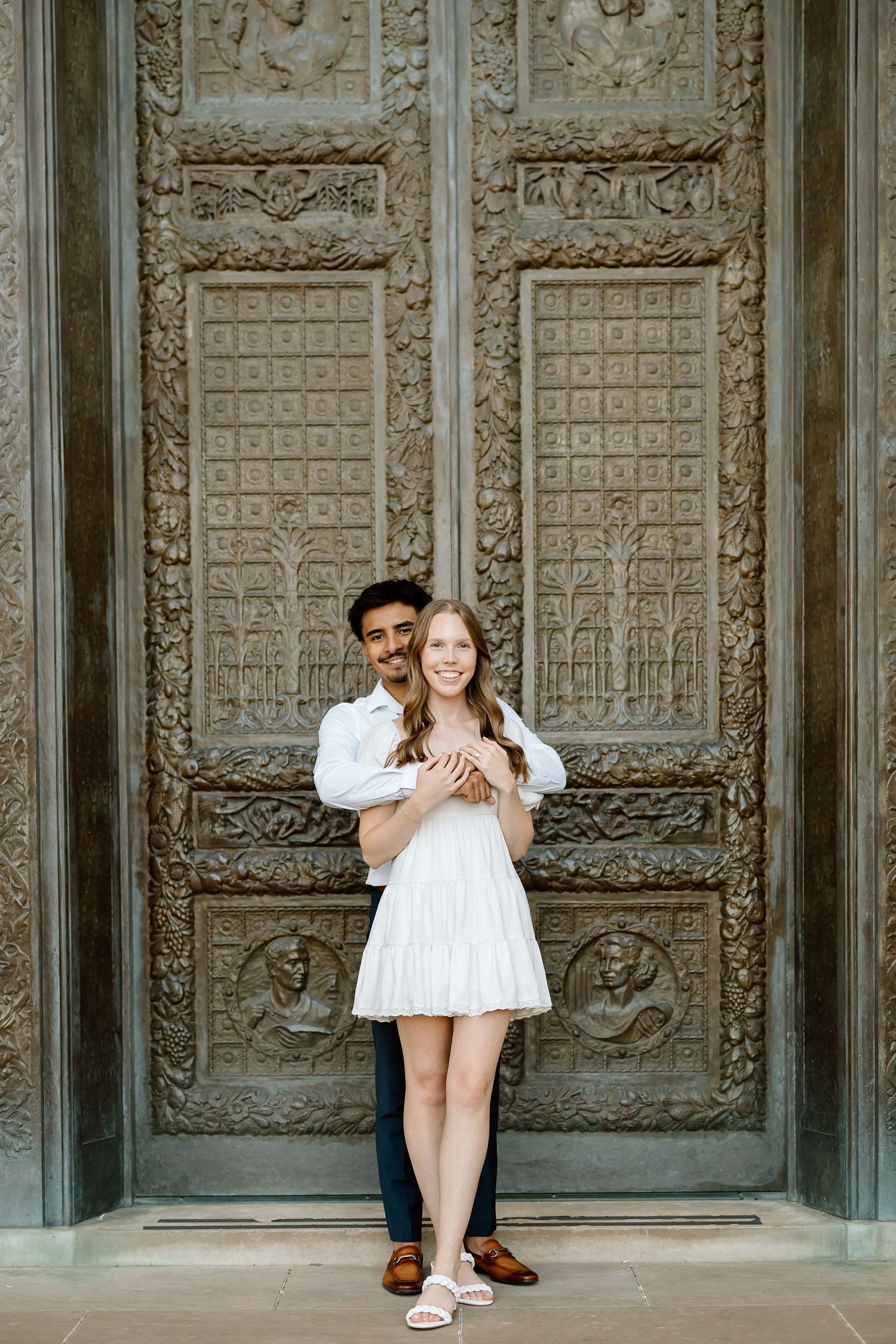 A couple poses in front of ornate carved metal doors with geometric patterns at a historic building entrance.