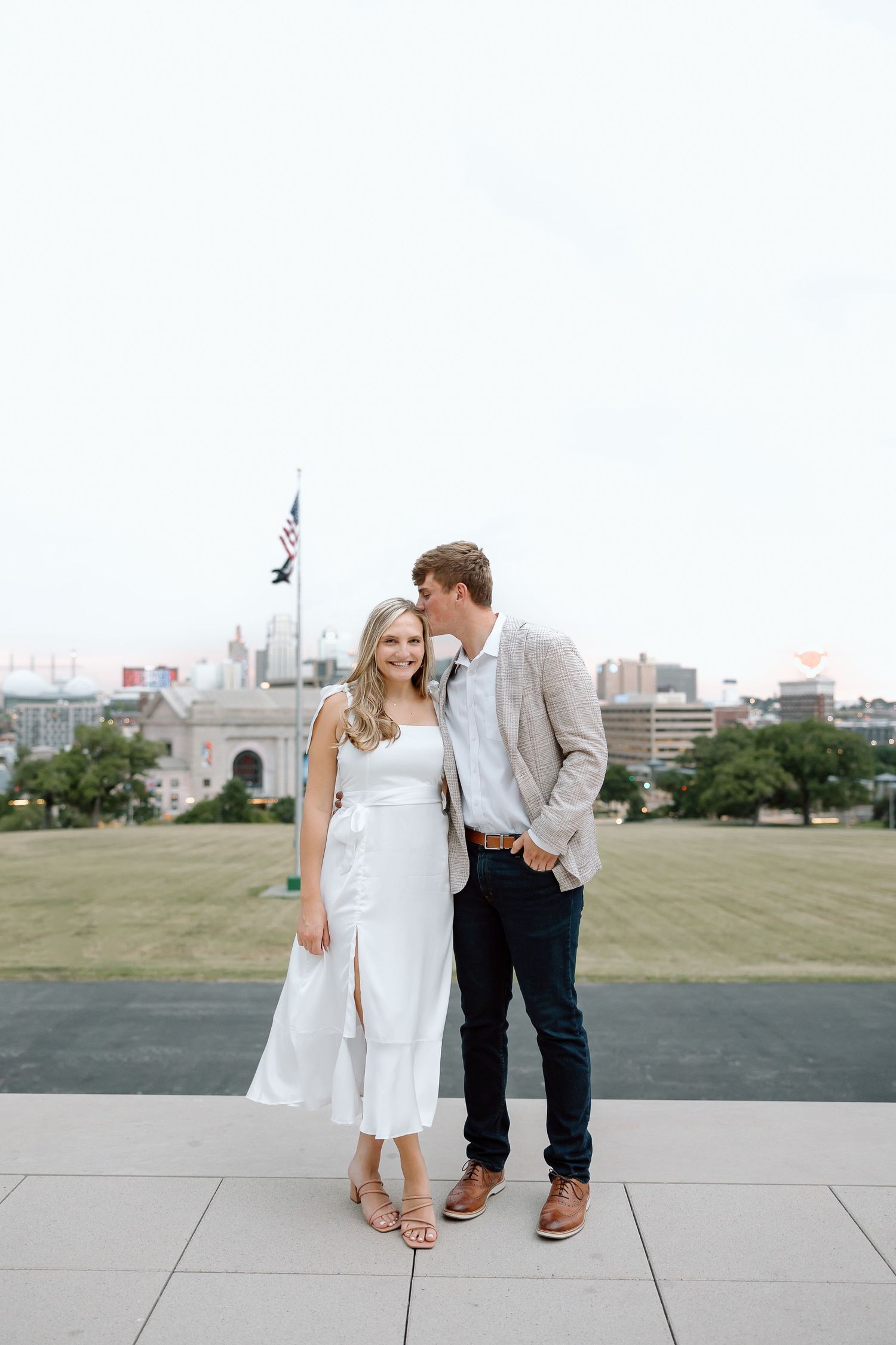 A couple shares a romantic moment on a concrete plaza with an urban skyline in the background.