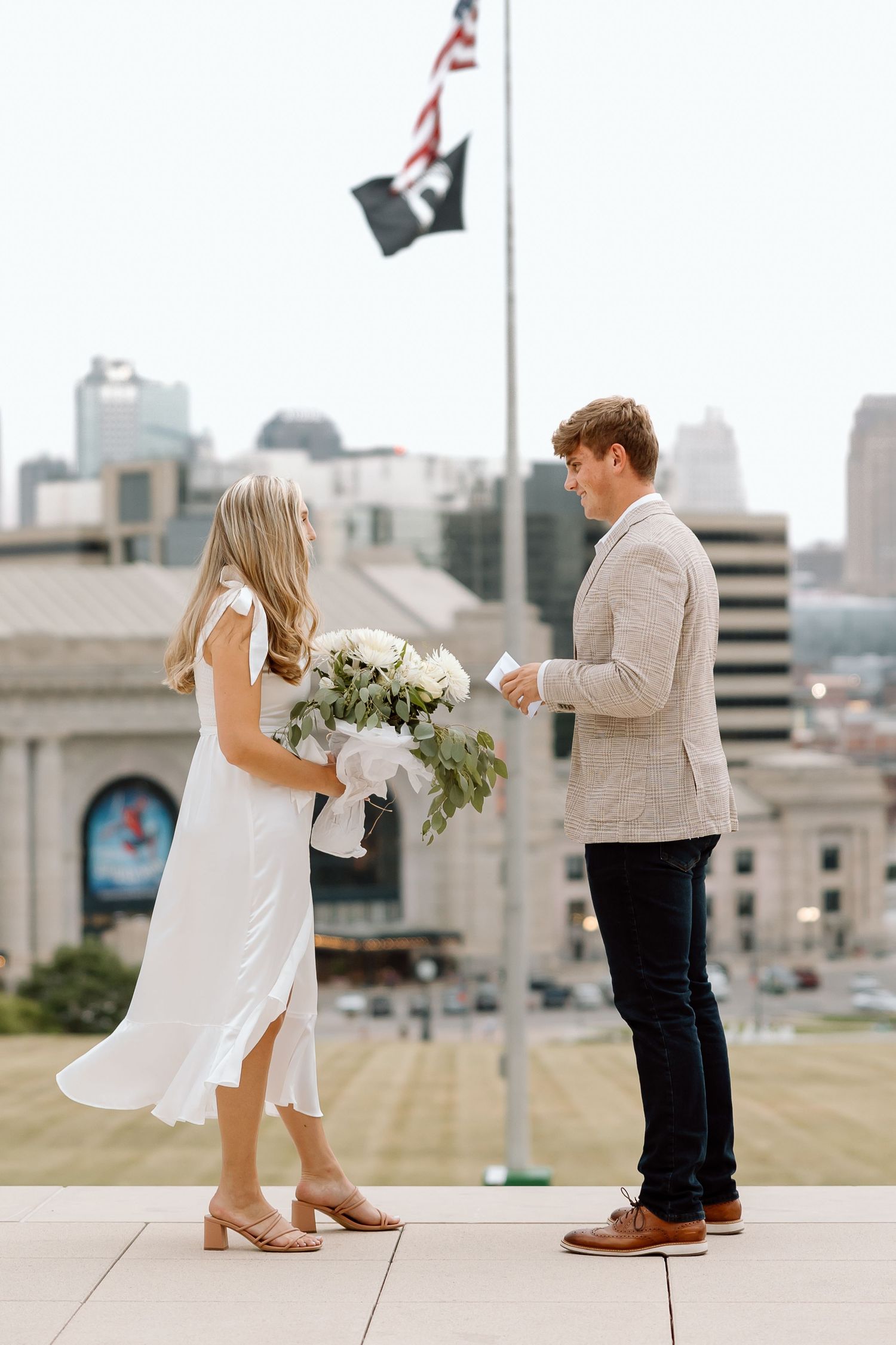 A romantic couple in white outfits dances together on a rooftop with a city skyline in the background.
