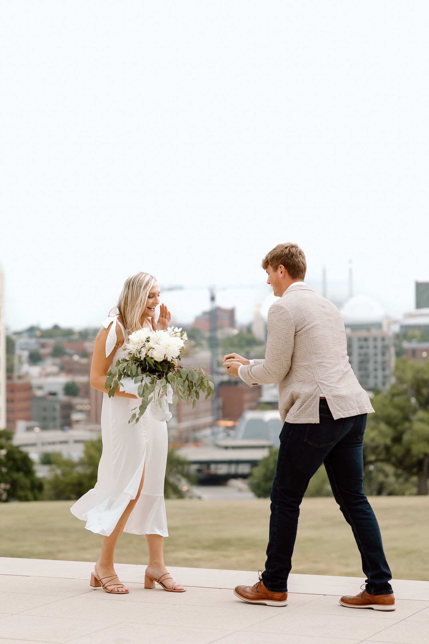 A couple in white outfits dance playfully together on a grassy hilltop overlooking a city skyline.