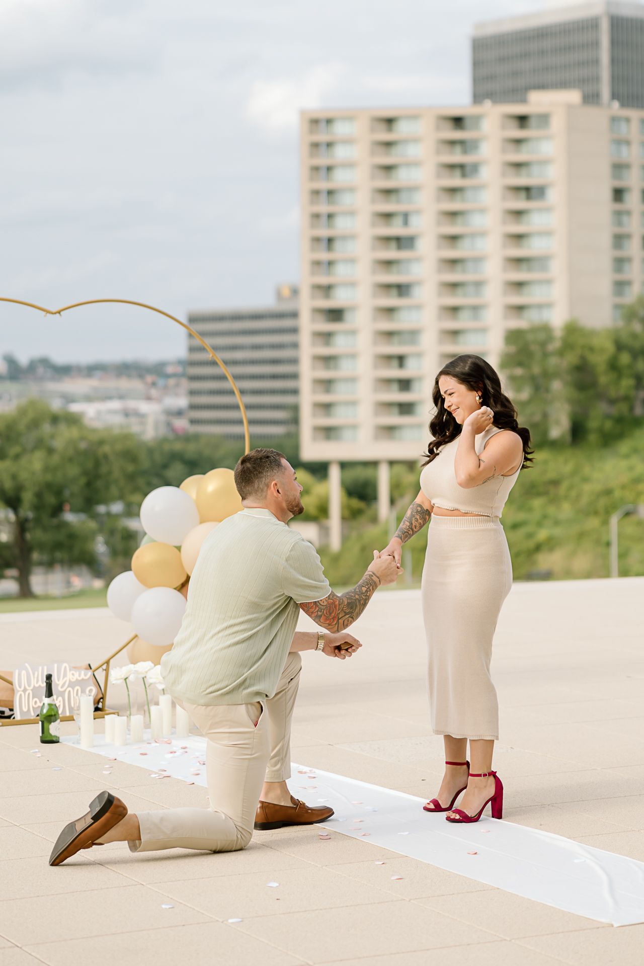 A couple sits on a white blanket with balloons and champagne on a rooftop terrace with city buildings in the background.