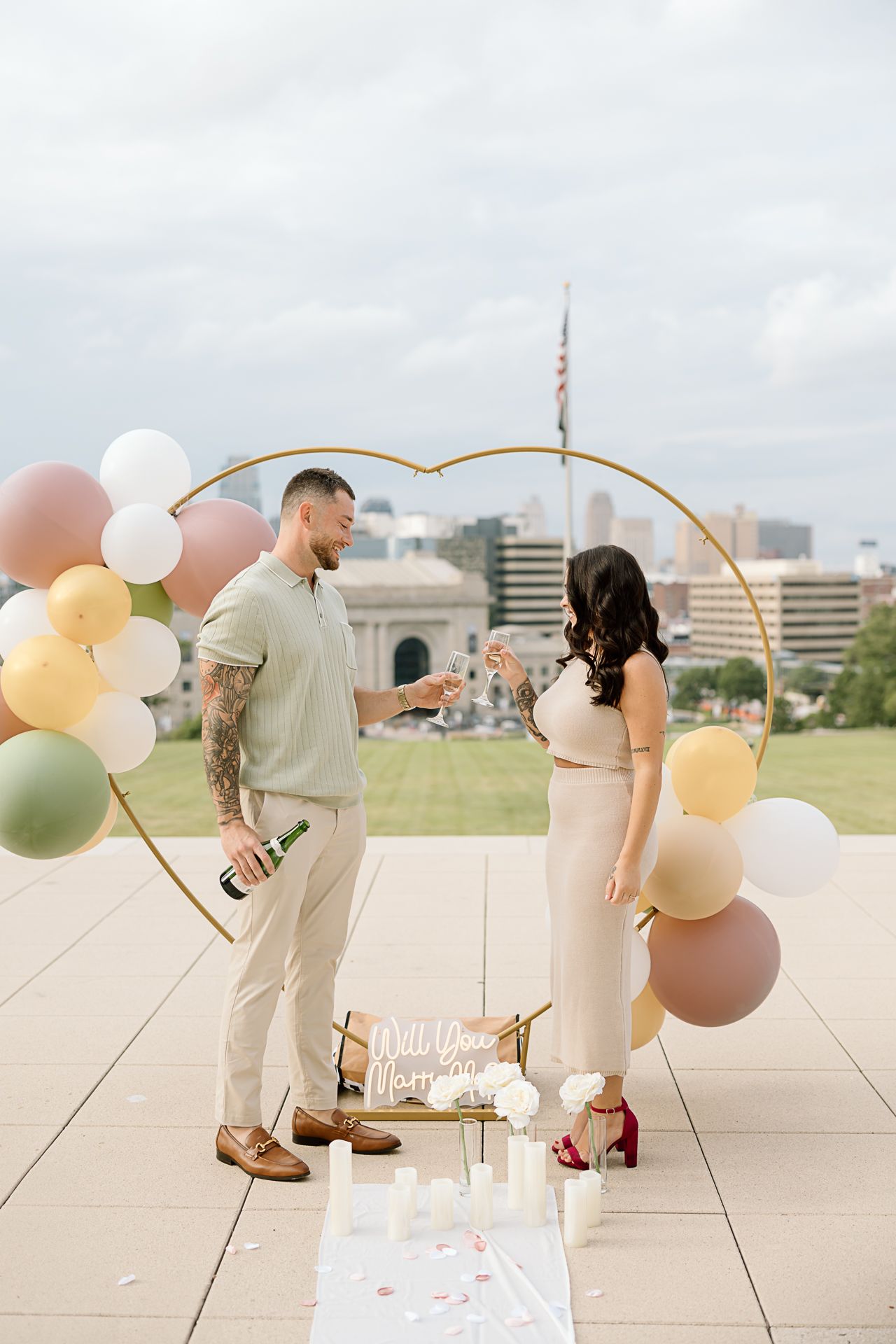 A romantic outdoor proposal setup with pastel balloons, champagne, and a decorative arch against a city skyline.