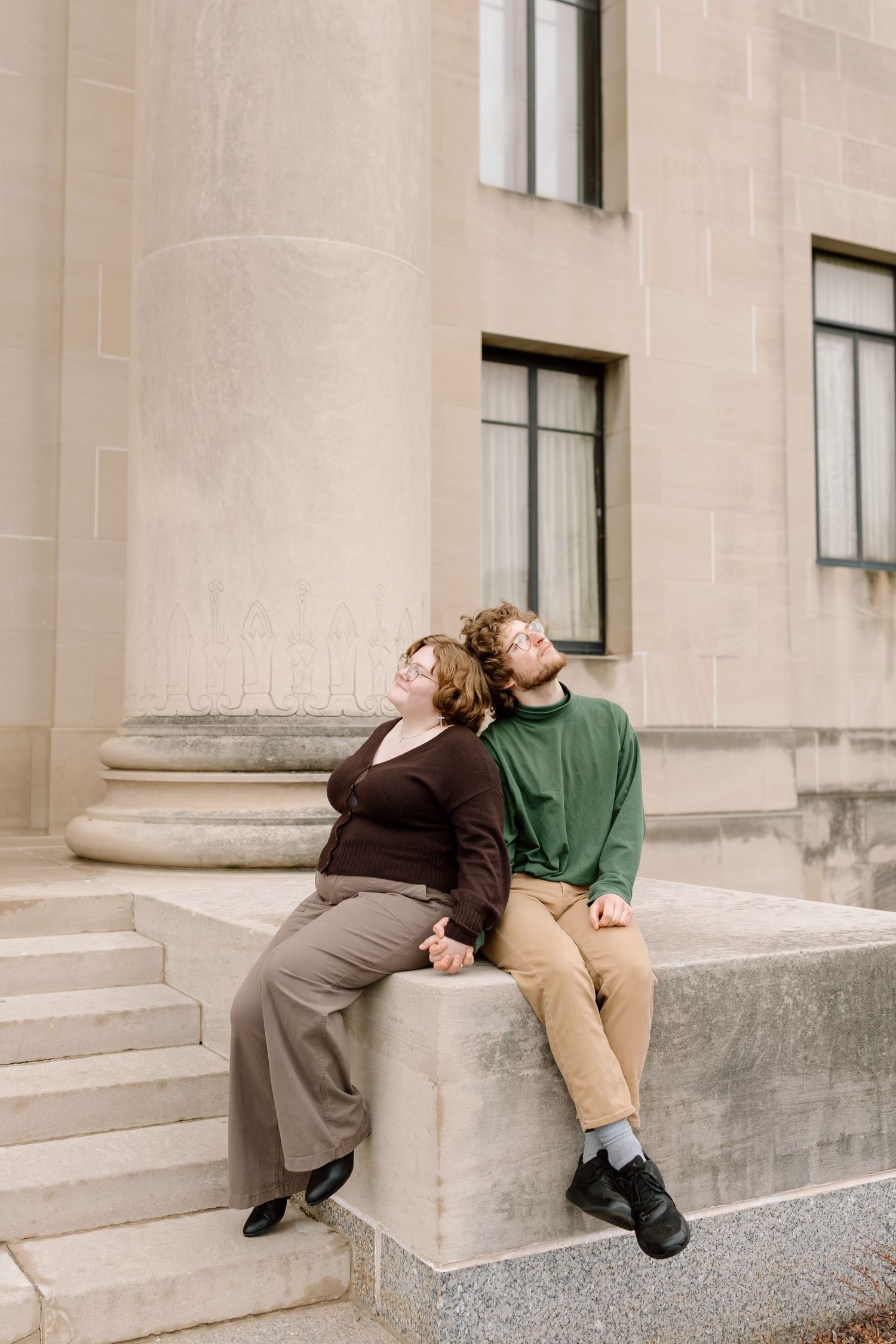 Two people in casual attire recline on stone steps against a beige building exterior.