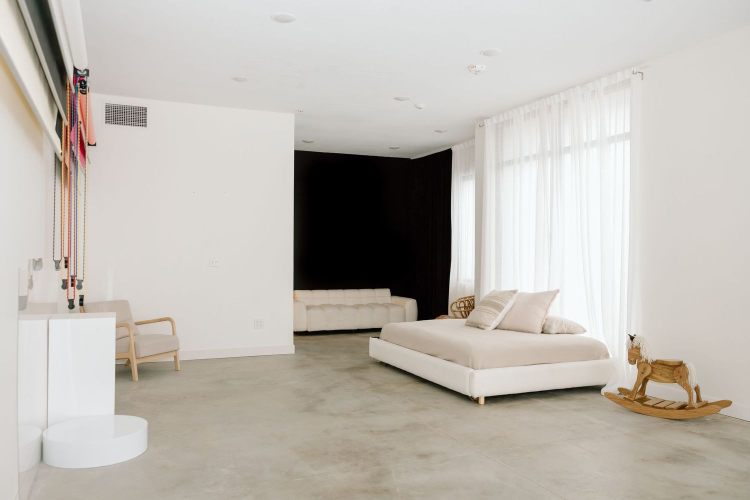 Modern minimalist bedroom interior with white walls, sheer curtains, and a wooden rocking horse by the window.