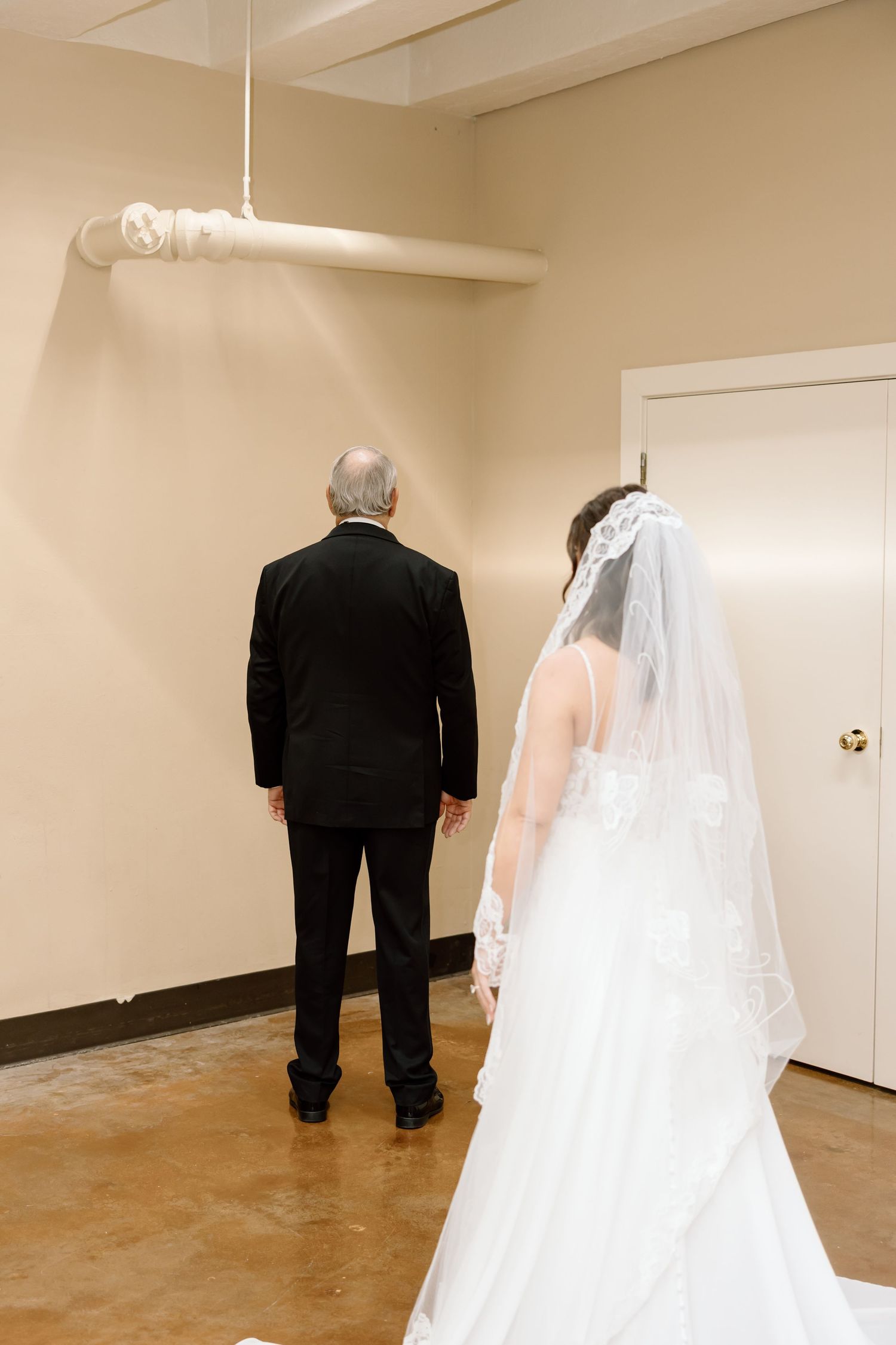 A bride in a white wedding dress and veil stands behind someone in a black suit in a simple hallway.