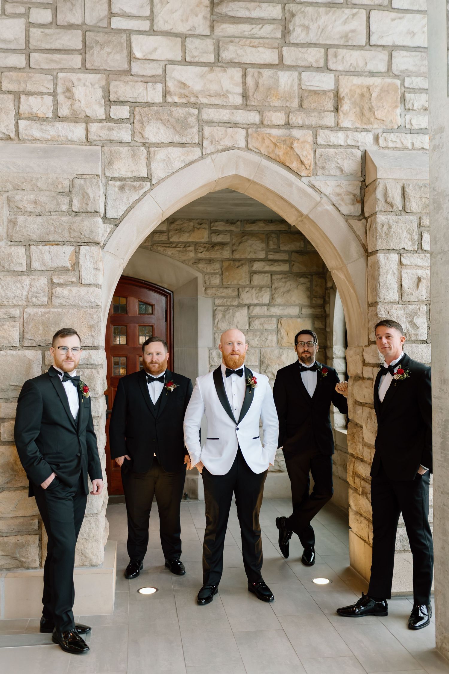 Groomsmen wearing tuxedos pose together under stone archways at a formal event.