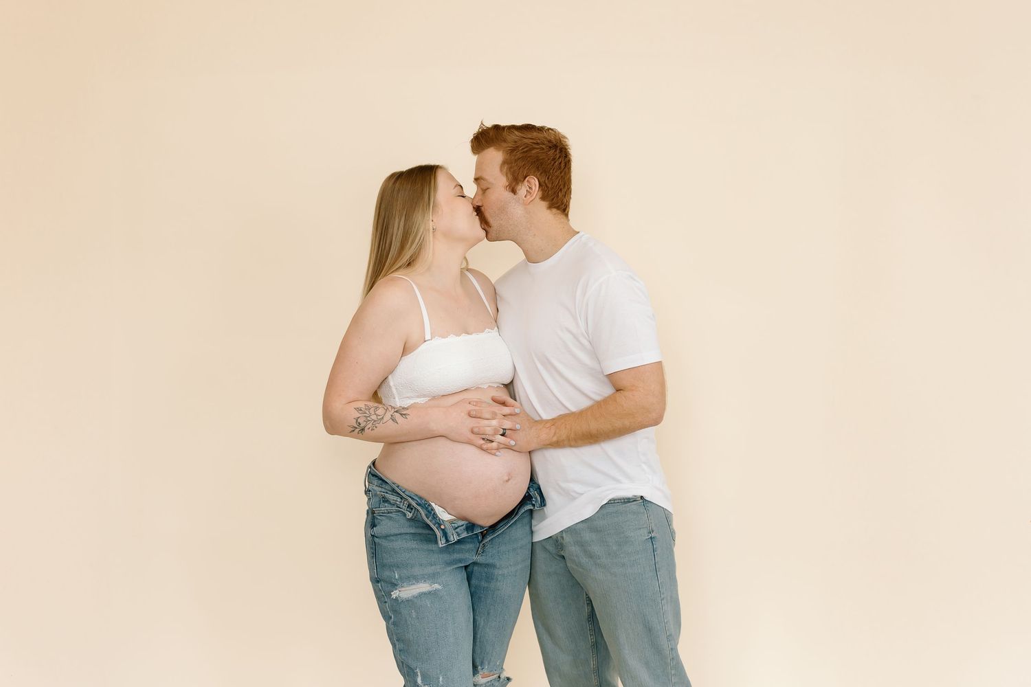 Candid maternity photo shows couple's natural affection against warm beige background.