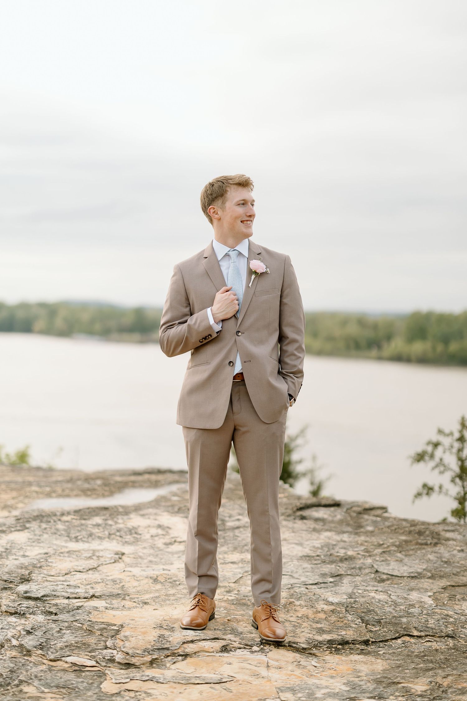 Groom in tan suit stands confidently on rocky overlook with water view.