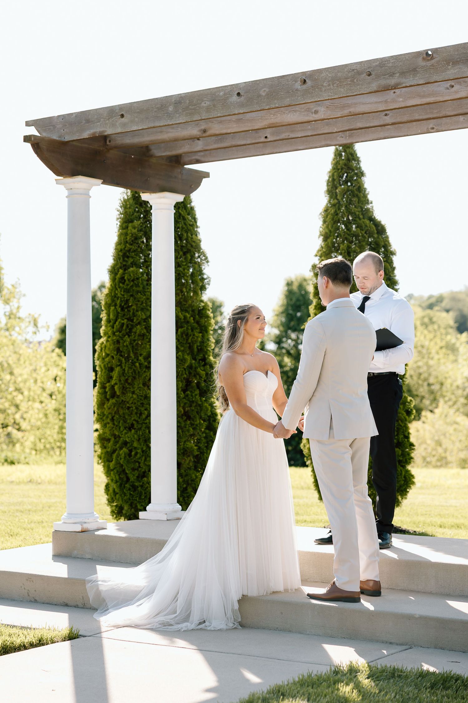 Wedding ceremony at white pergola with tall cypress trees framing the elegant outdoor setting.