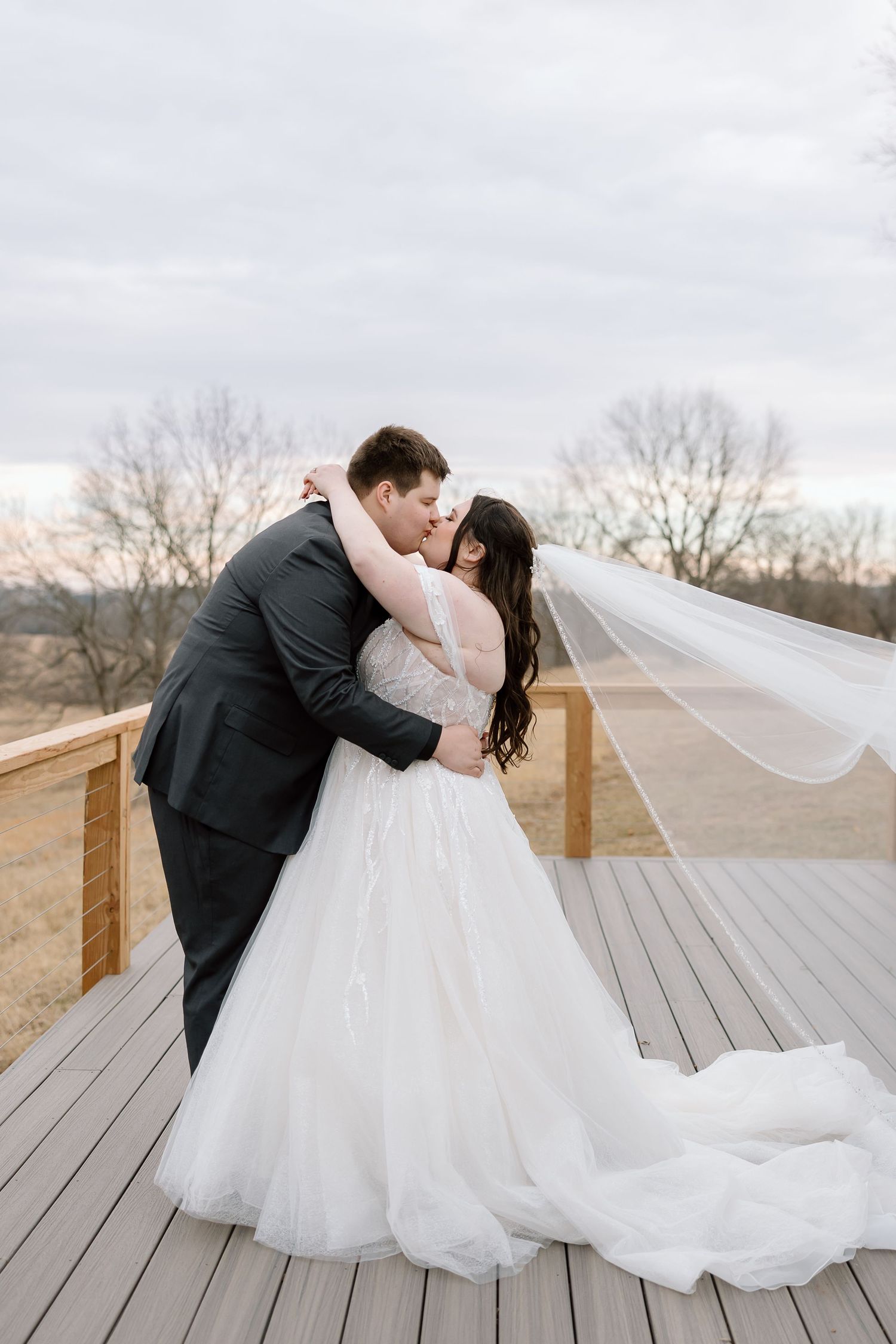 Couple shares romantic moment on wooden deck with flowing veil and winter landscape.