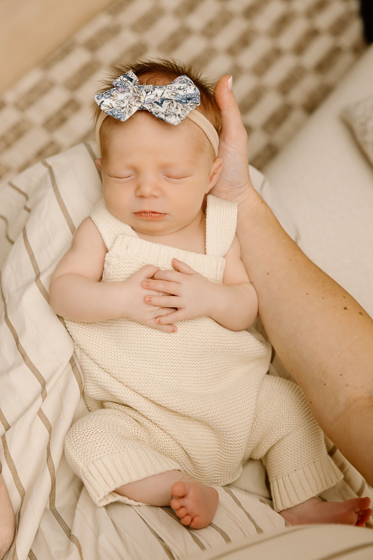 Sweet newborn in cream romper and sparkly bow resting on textured cushions.