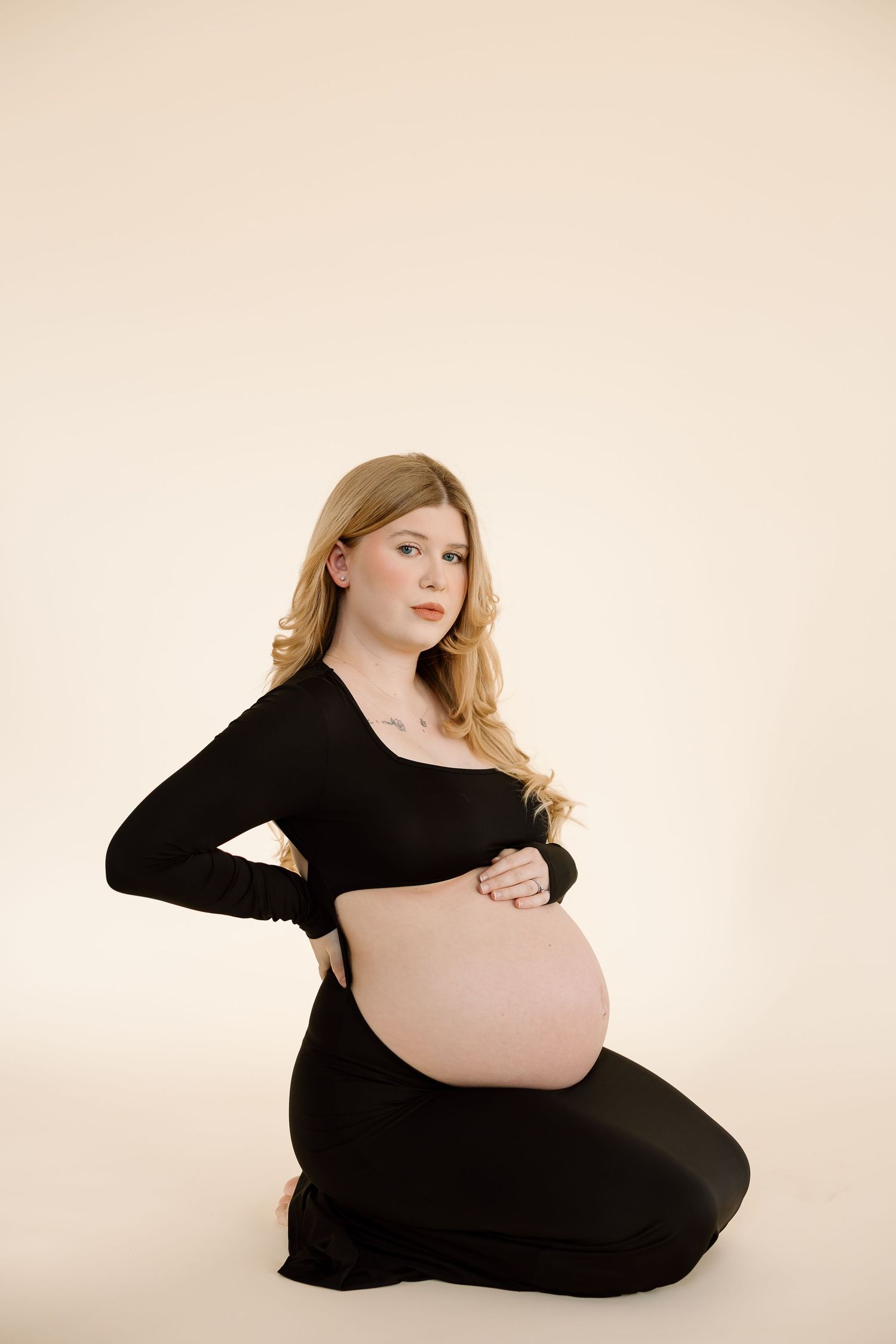 Side view maternity photo featuring pregnant woman in long-sleeved black dress on neutral backdrop.