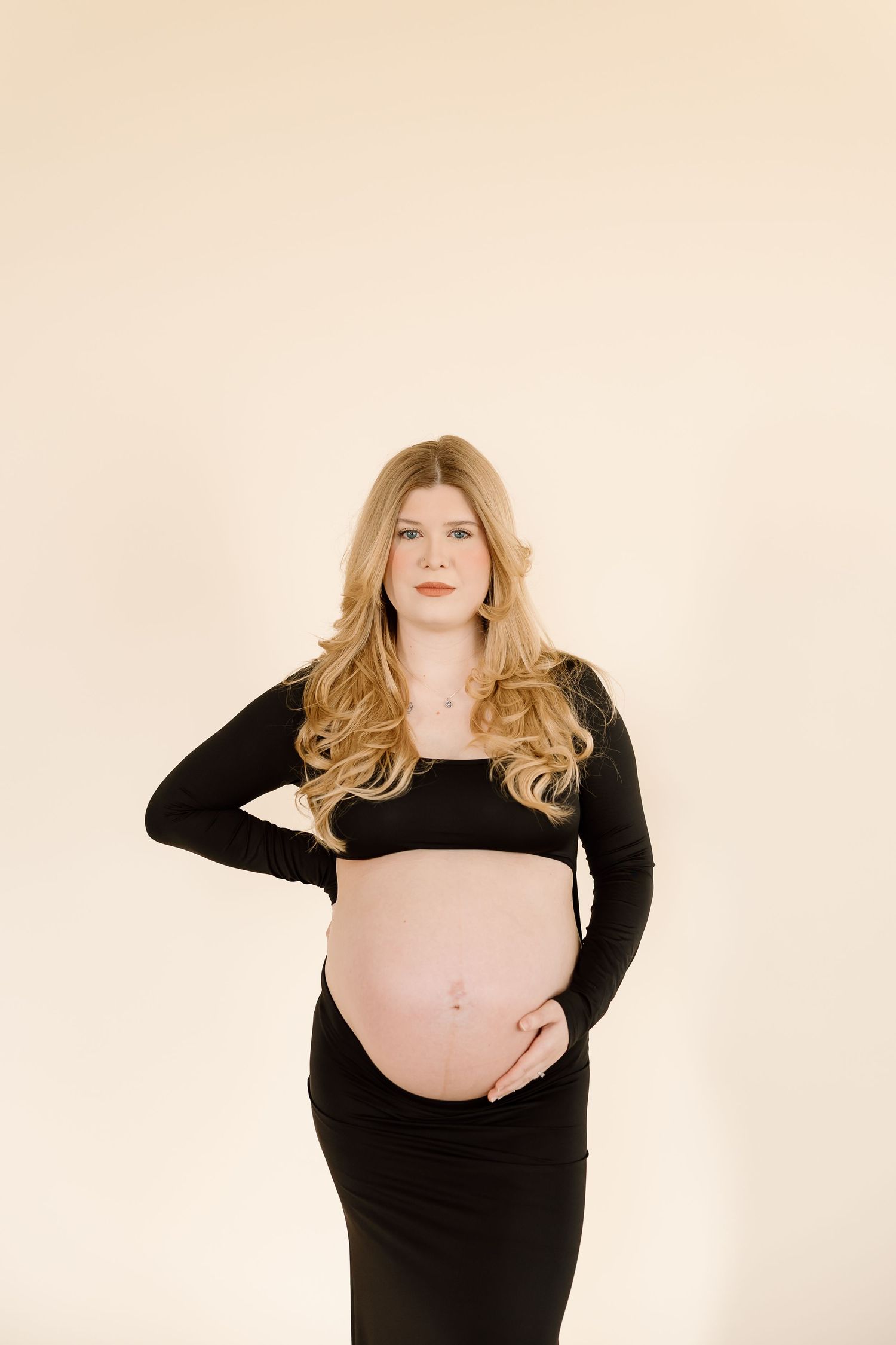 Modern maternity photo with woman in black crop top and skirt against beige background.