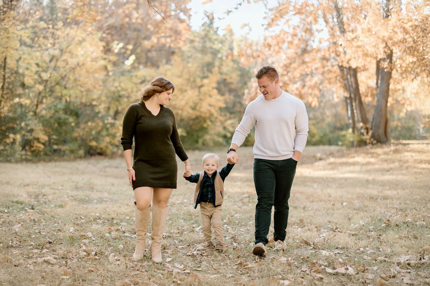 Family of three walks together holding hands in a fall setting with golden leaves.