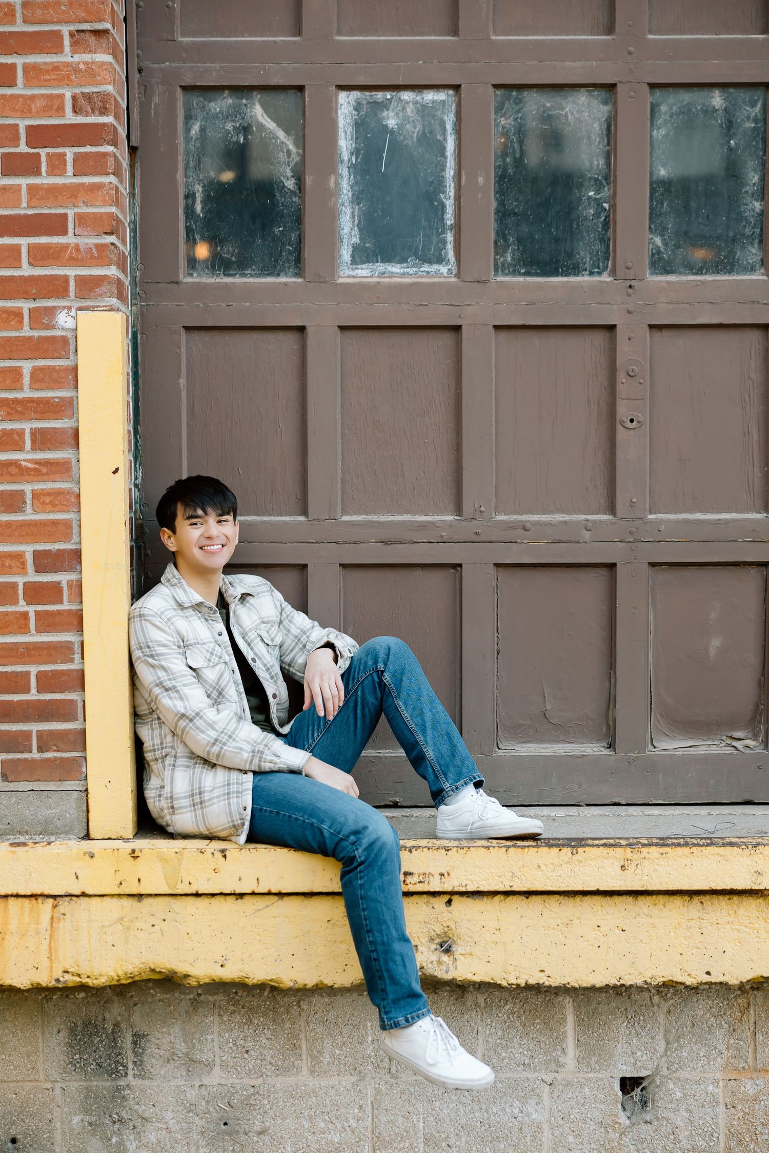 Young person in plaid jacket and jeans sitting casually against brown door with vintage glass panels.