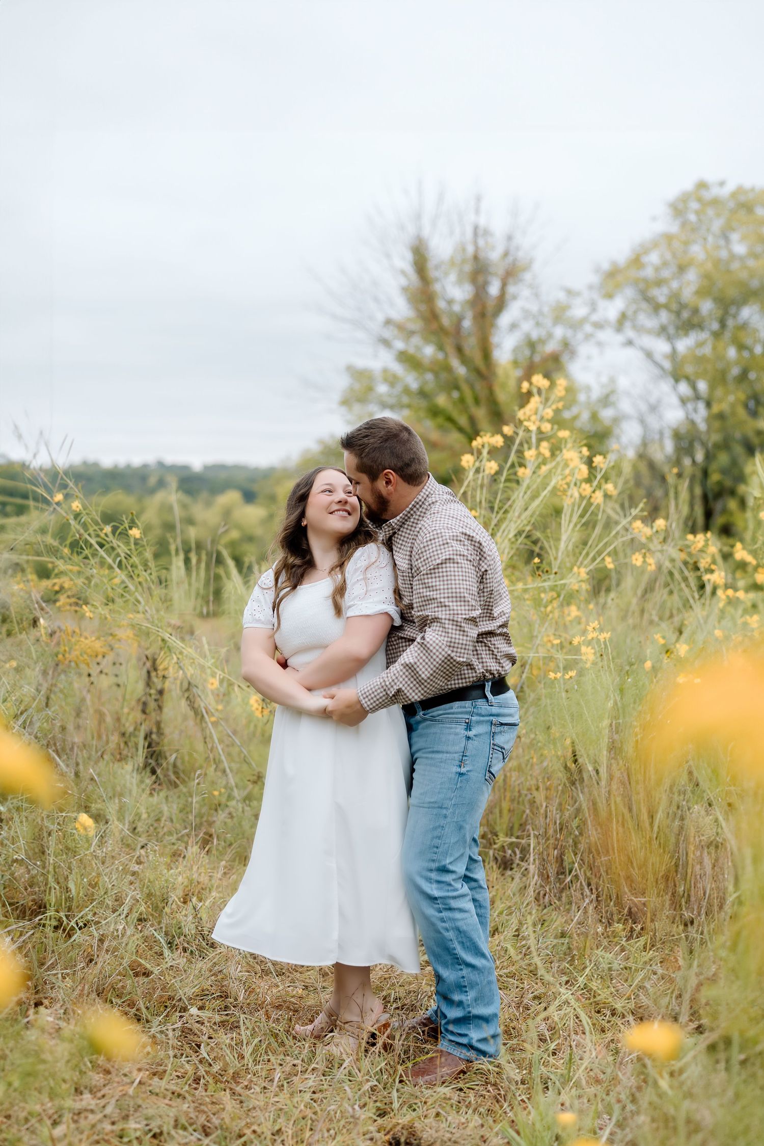 A couple in casual attire embraces in a rustic outdoor setting with yellow flowers.