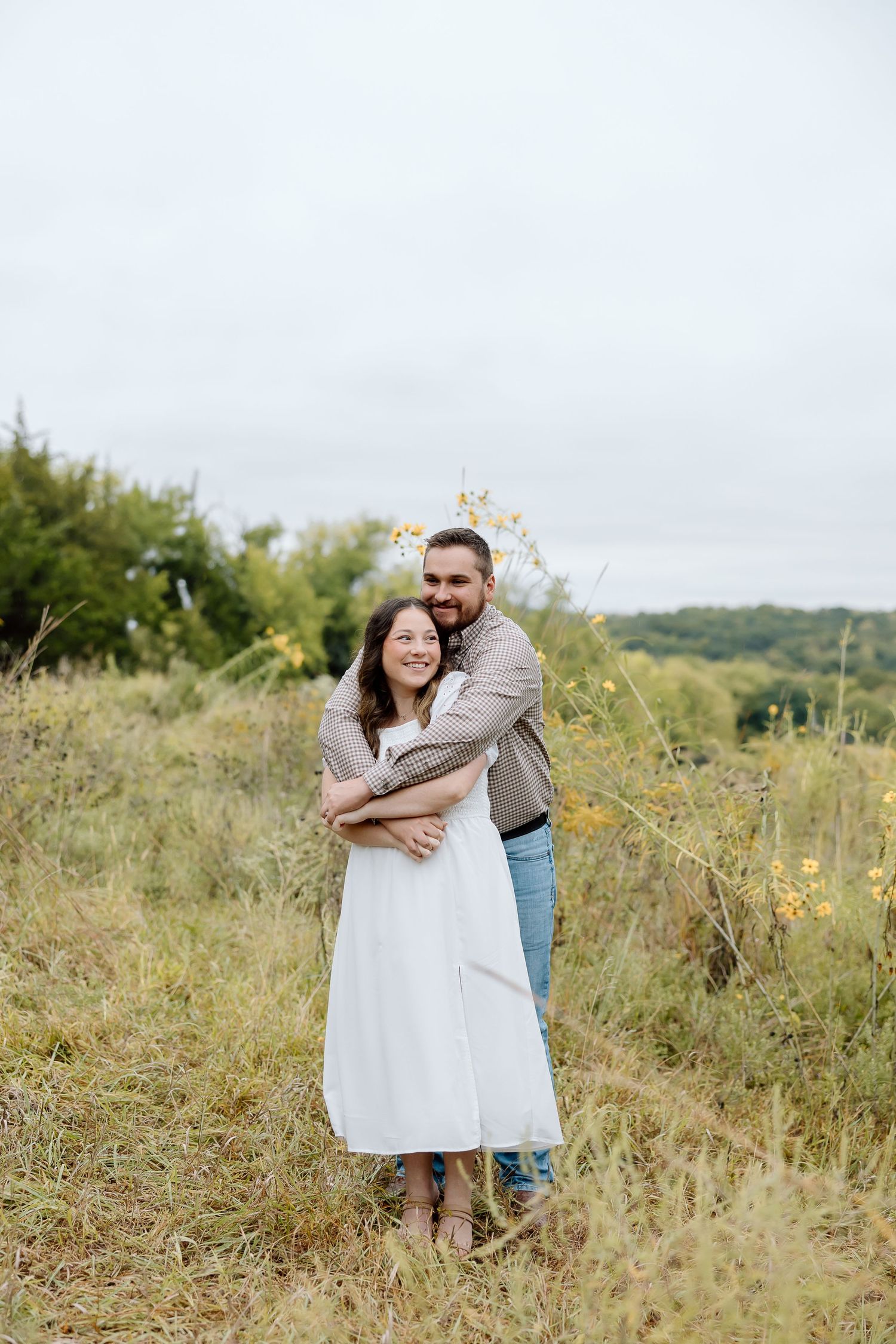 A romantic couple embraces in a field of tall grass on an overcast day.