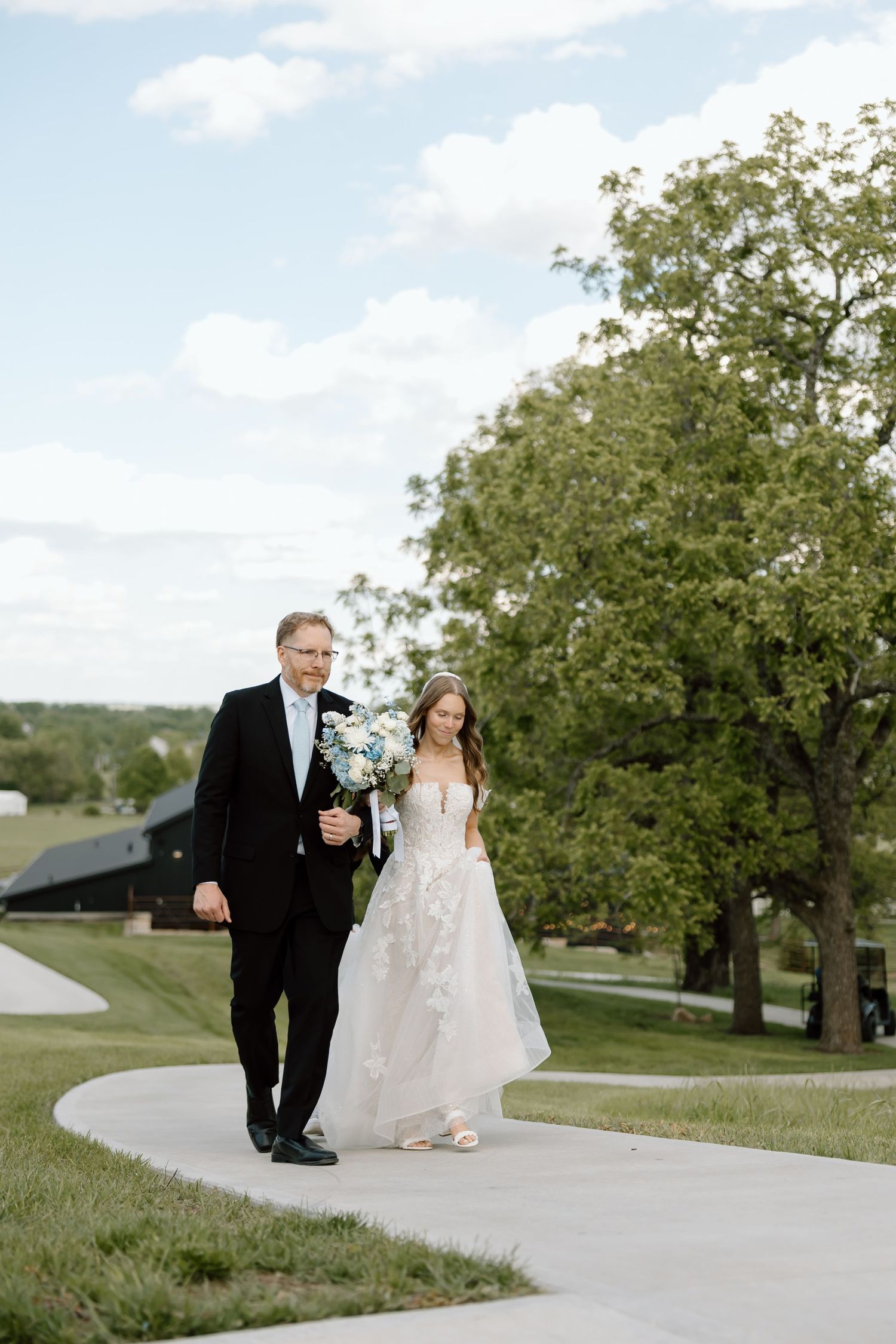 Bride in lace gown walks with father in tuxedo along curved path, holding blue and white bouquet on wedding day.