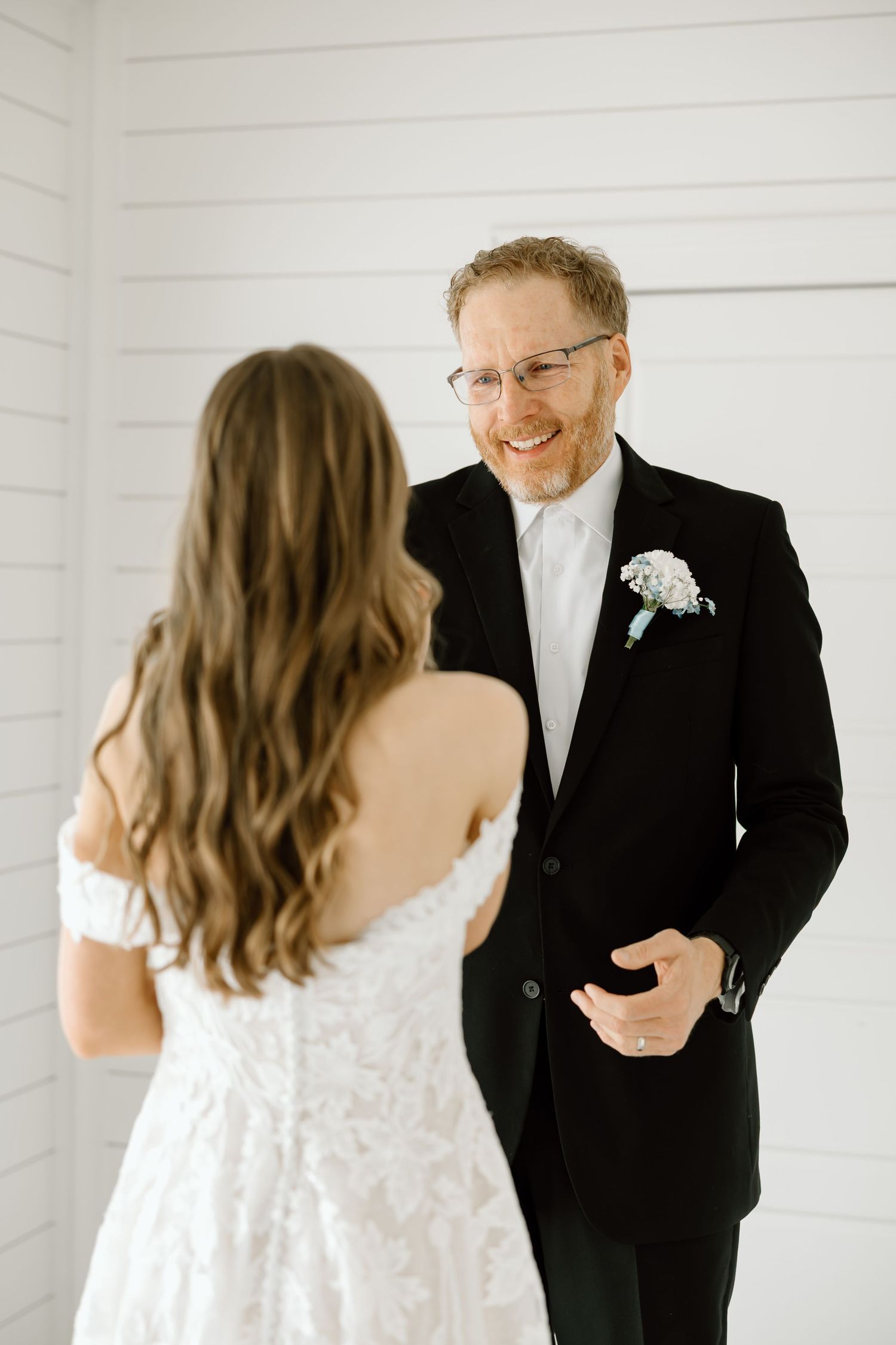Smiling groom in black tuxedo with boutonniere shares a first look moment with bride in white off-shoulder gown.