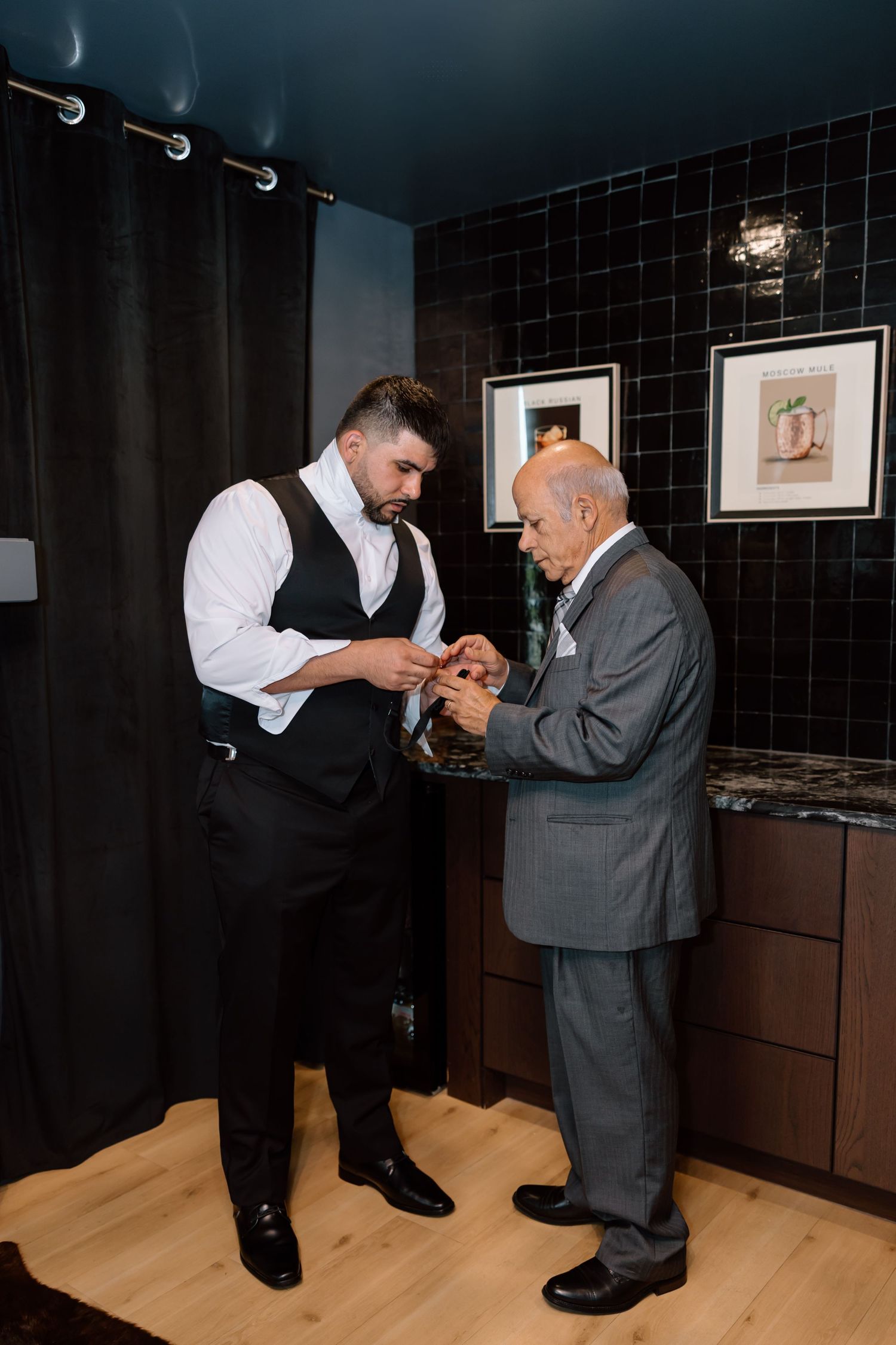 Groom in vest and older man in gray suit sharing a moment while adjusting cufflinks in a modern black-tiled room.