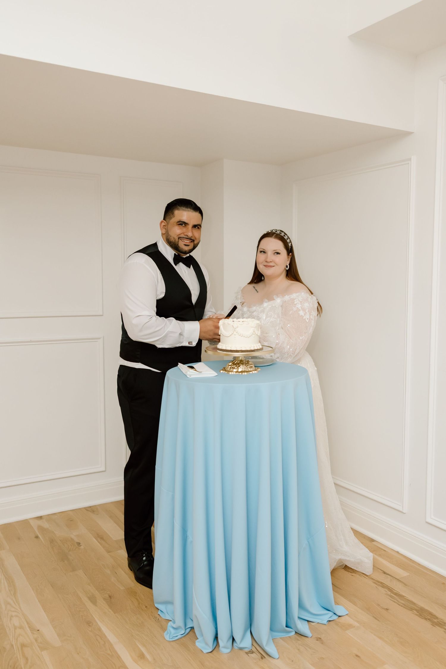 Bride and groom cutting their white wedding cake together at a blue draped table in a bright white venue.