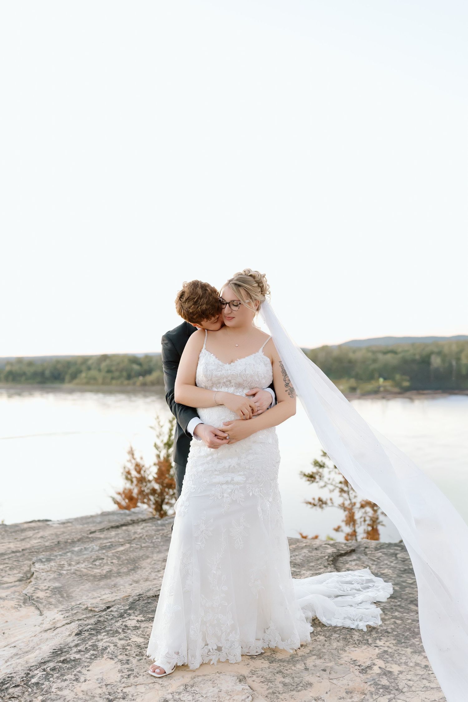 A romantic wedding couple embraces on a sandy beach during golden hour with a flowing white veil and lace dress.