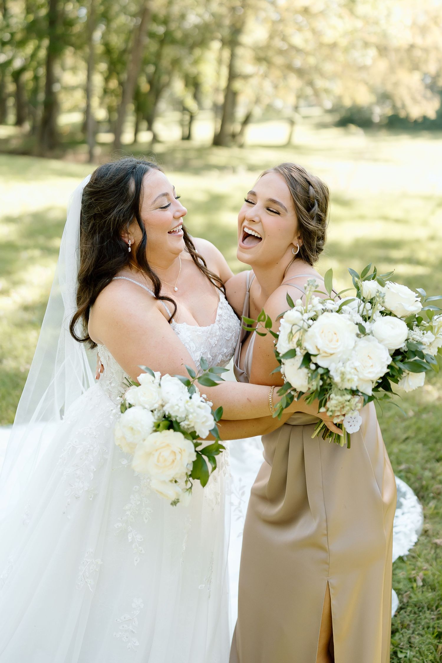 Two people in wedding attire share joyful laughter while holding white floral bouquets in an outdoor garden setting.