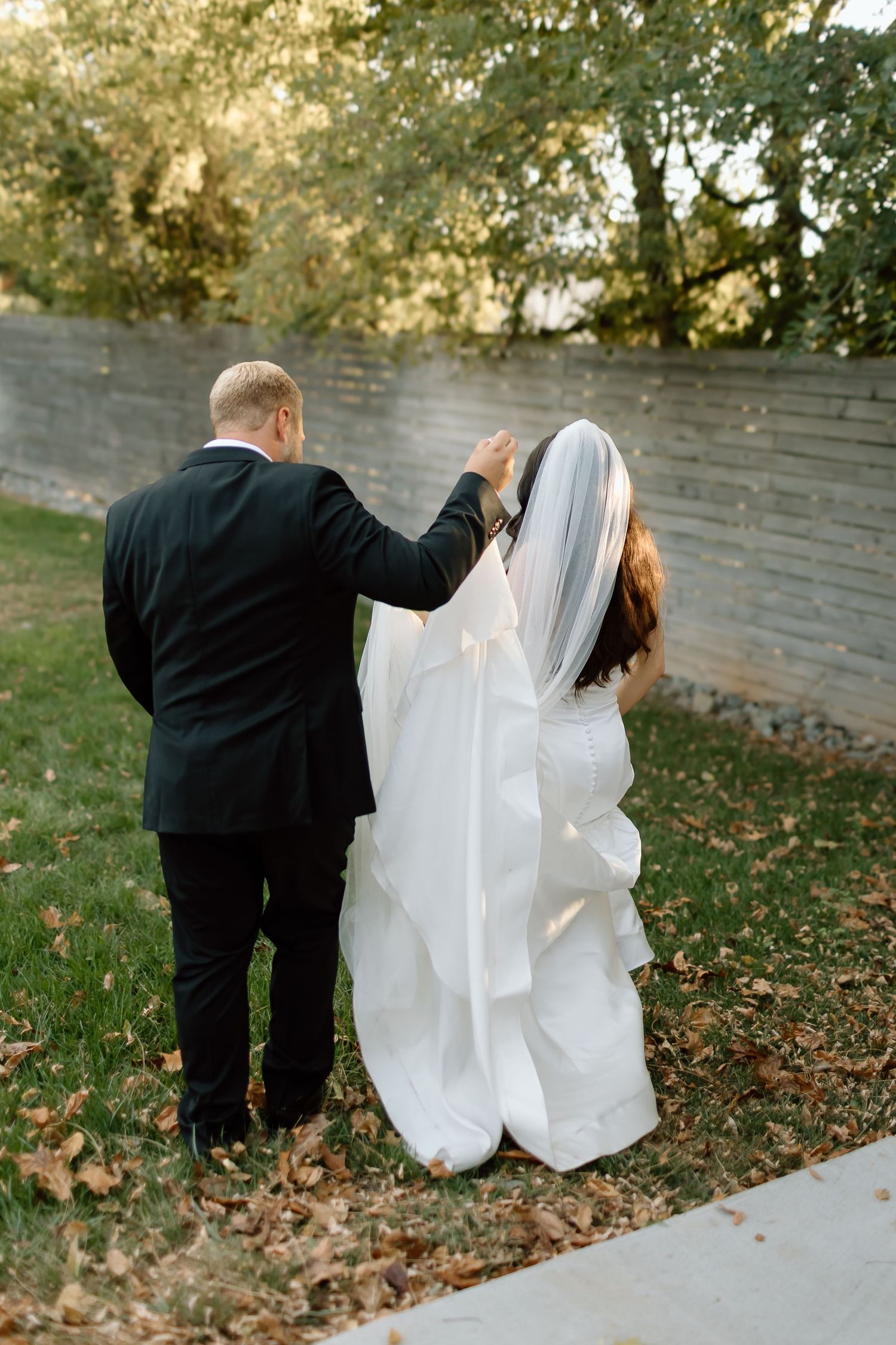A couple in wedding attire walks along a garden path on their special day, with autumn leaves scattered on the ground.