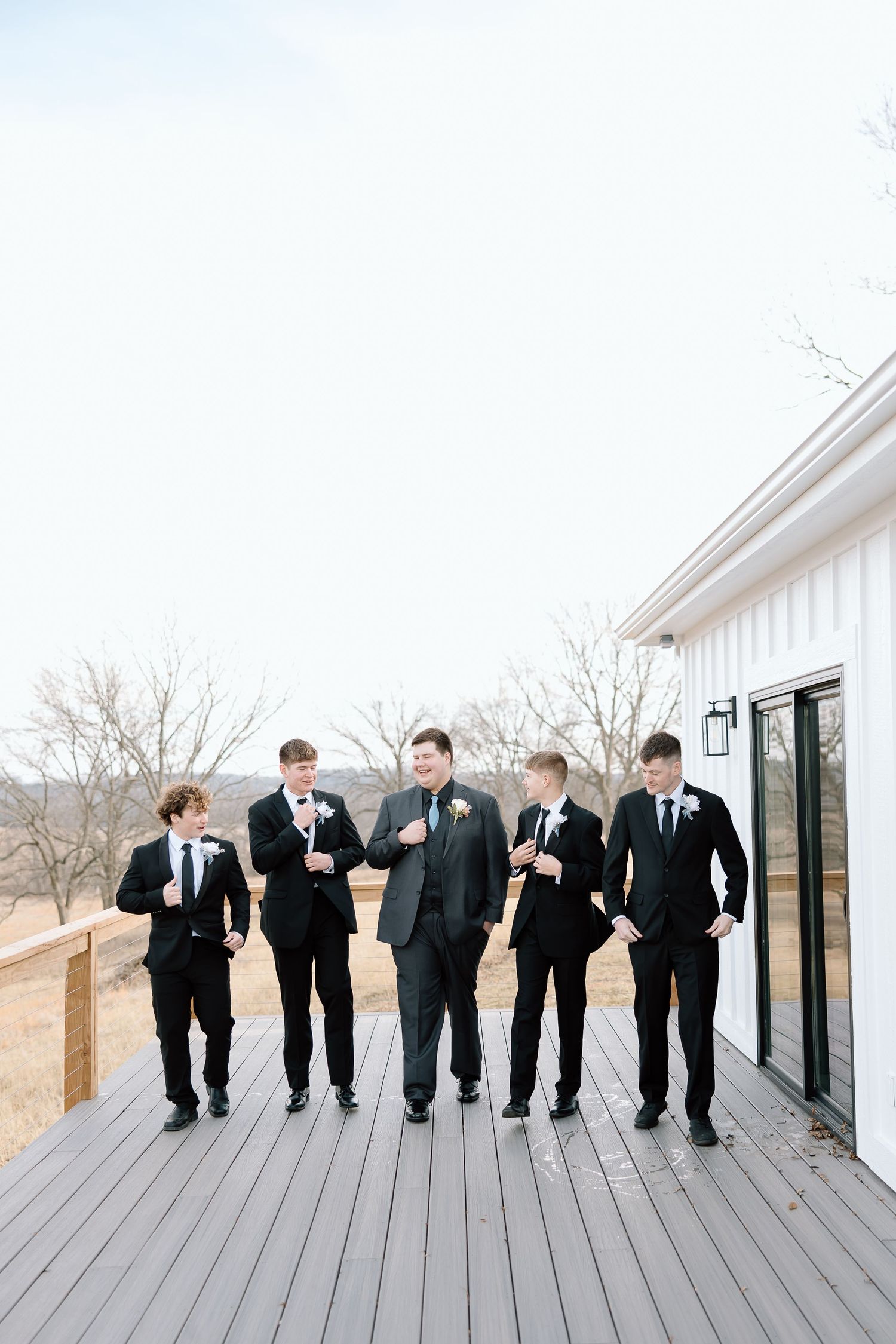 A group of groomsmen in black suits walk and laugh together on a modern wooden deck.