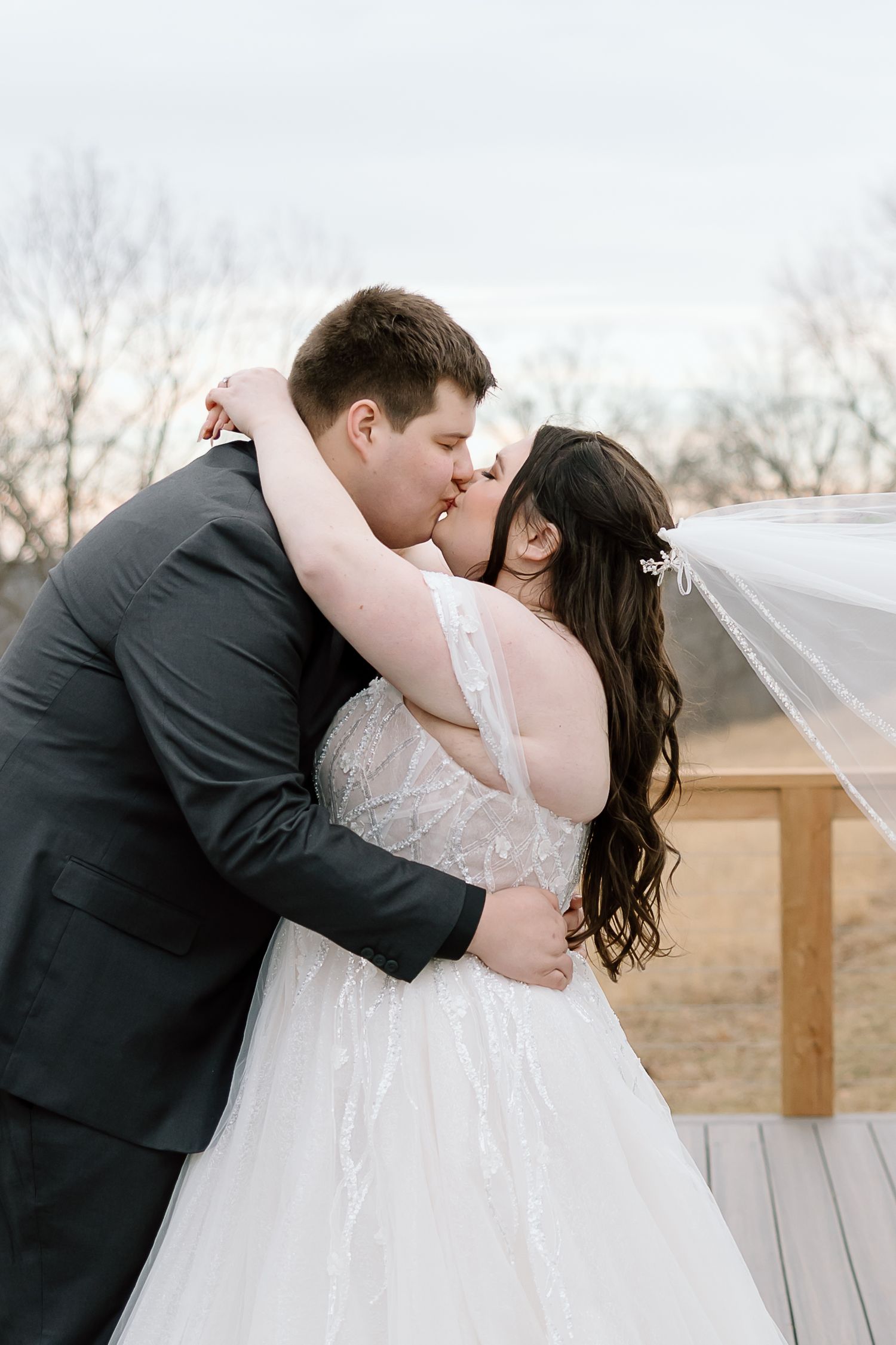A romantic wedding moment captured on a deck as newlyweds share a tender kiss under a cloudy winter sky.