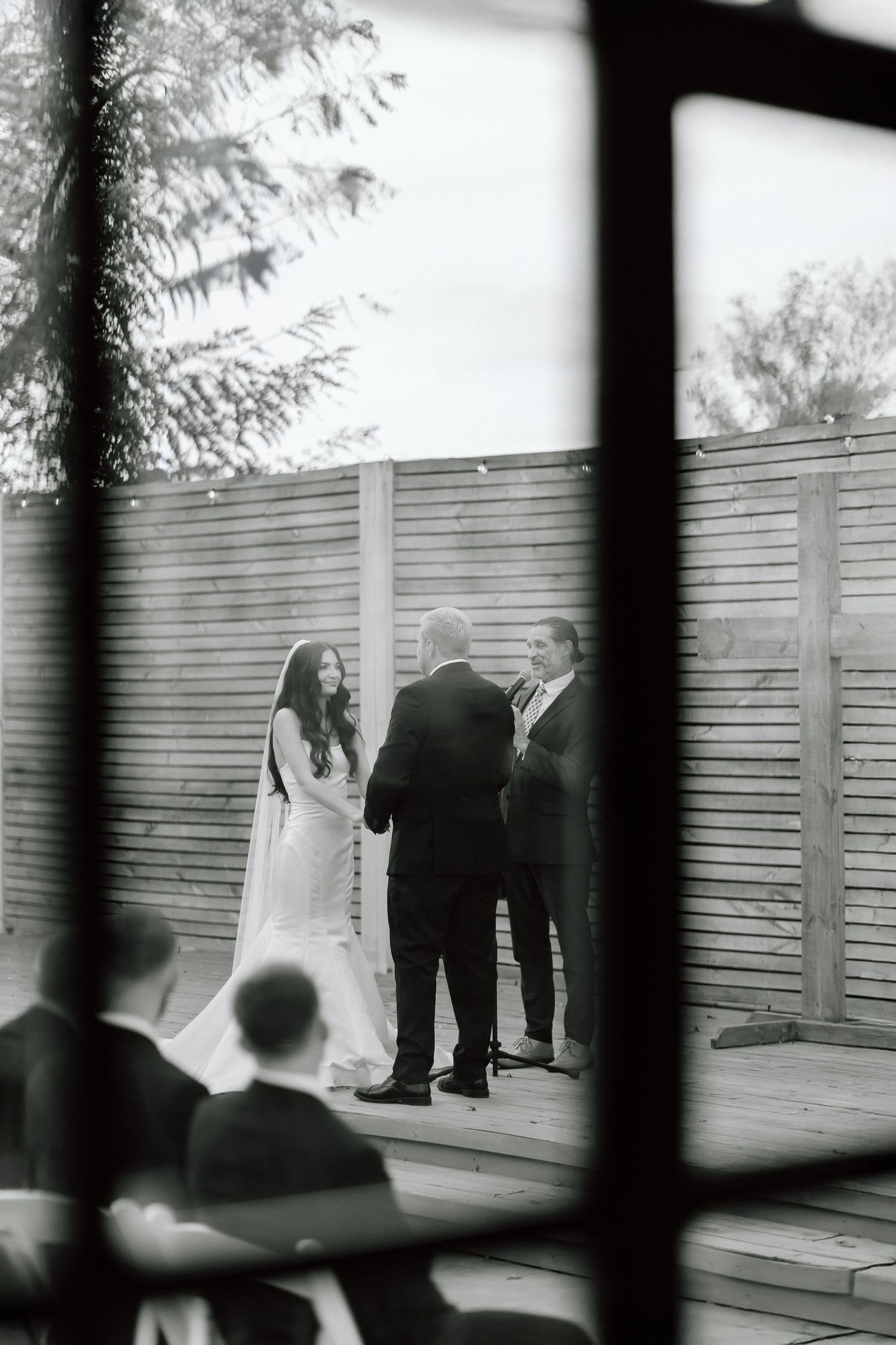 A black and white wedding ceremony photo captured through a window frame showing a couple at the altar.