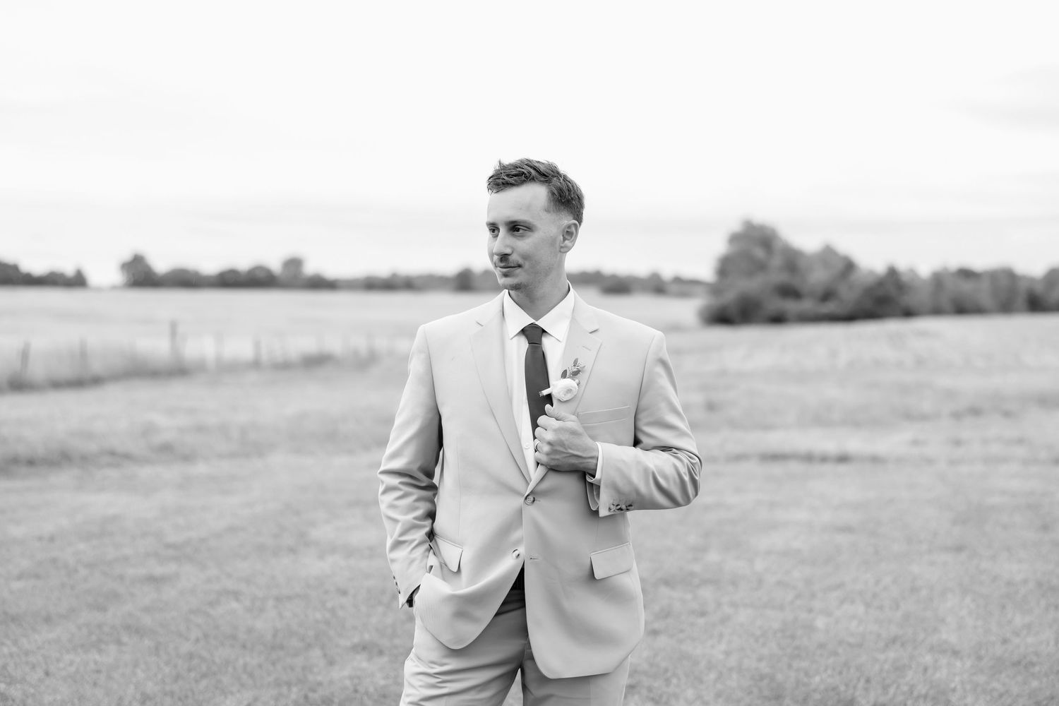 Black and white photo of a groom in a light suit standing in a field, looking away confidently.
