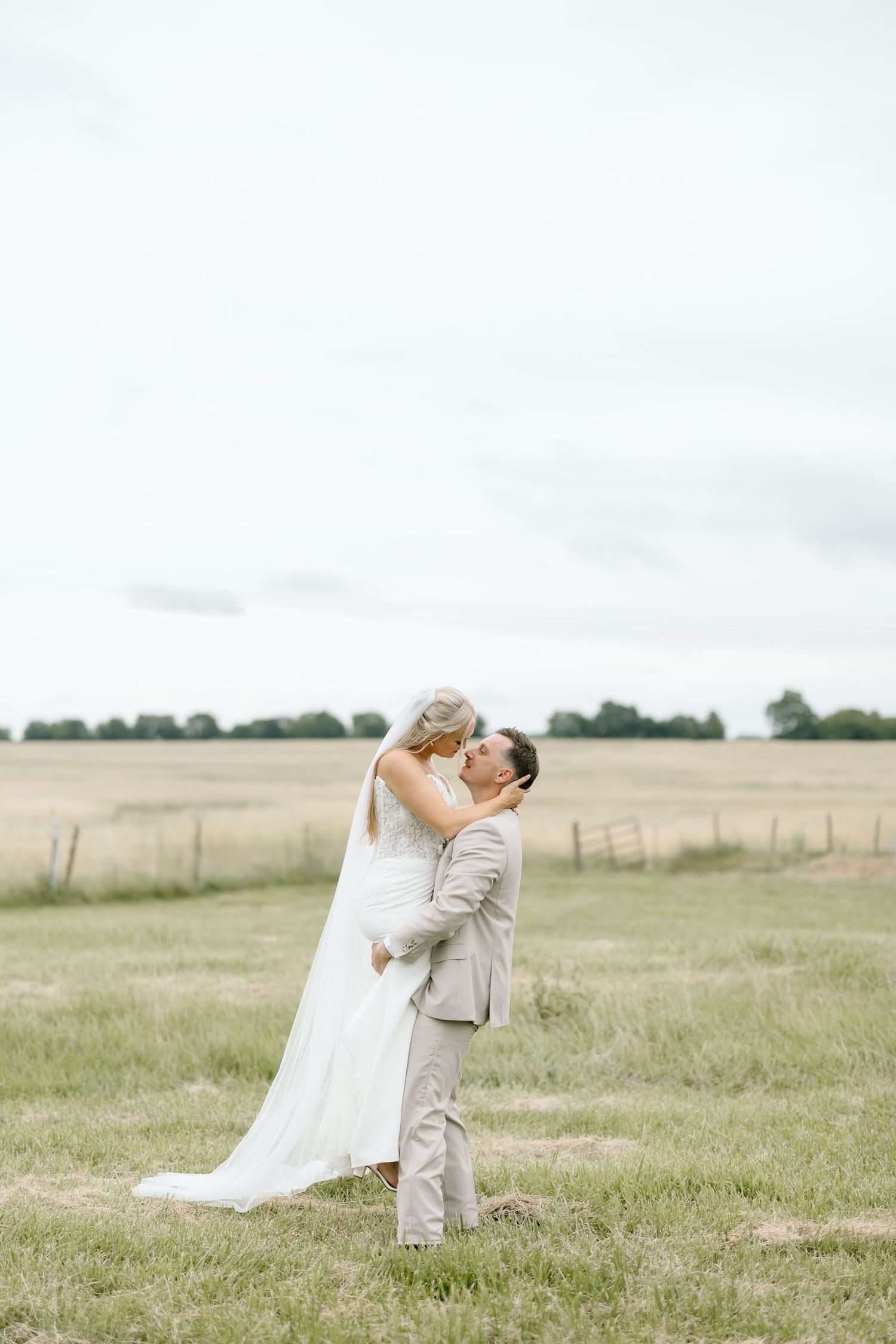 Groom lifting bride in white gown and veil in a scenic countryside field on their wedding day.