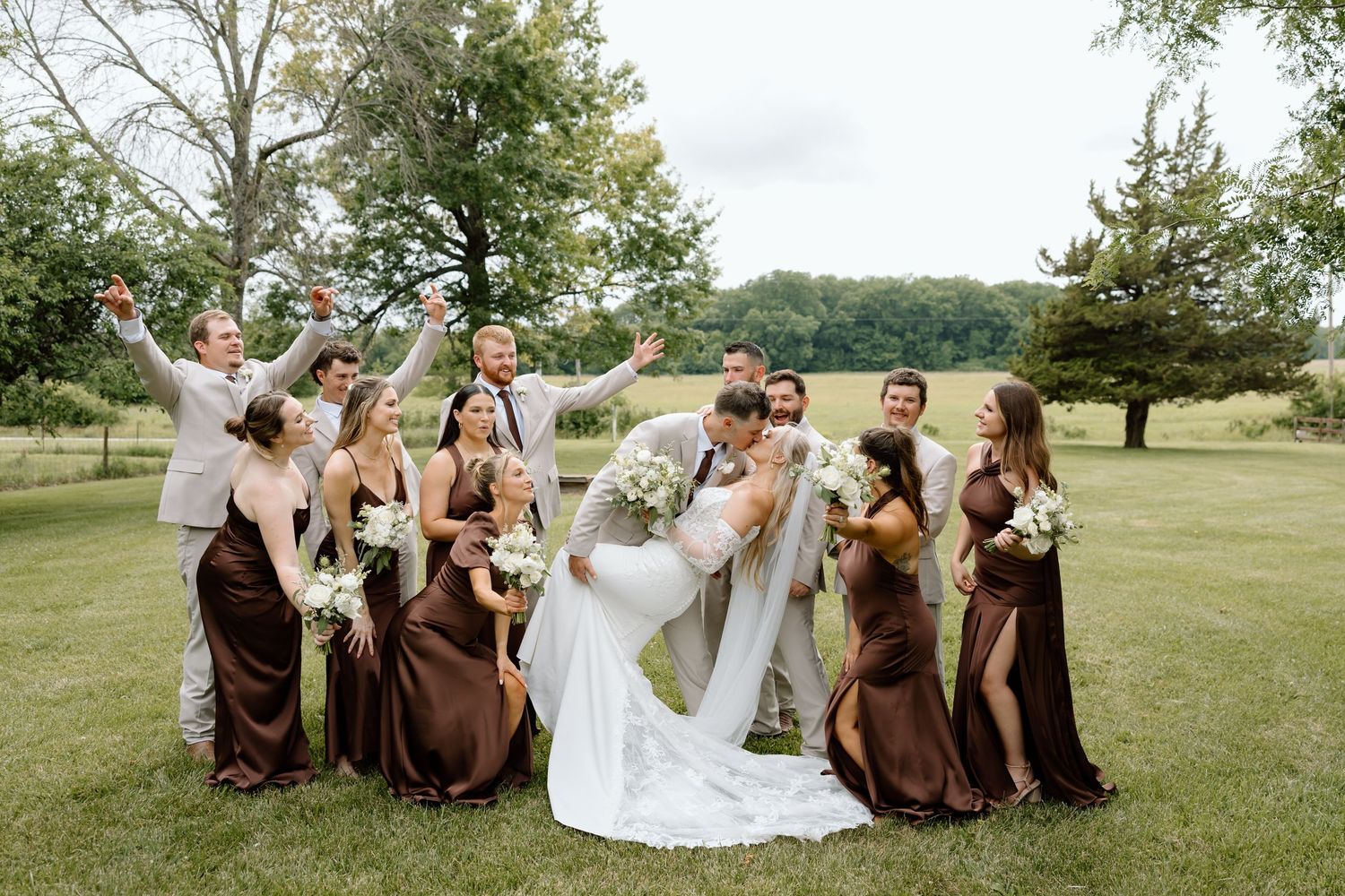 Wedding party celebrates outdoors as bride and groom kiss, bridesmaids in brown dresses and groomsmen in gray suits on green lawn.