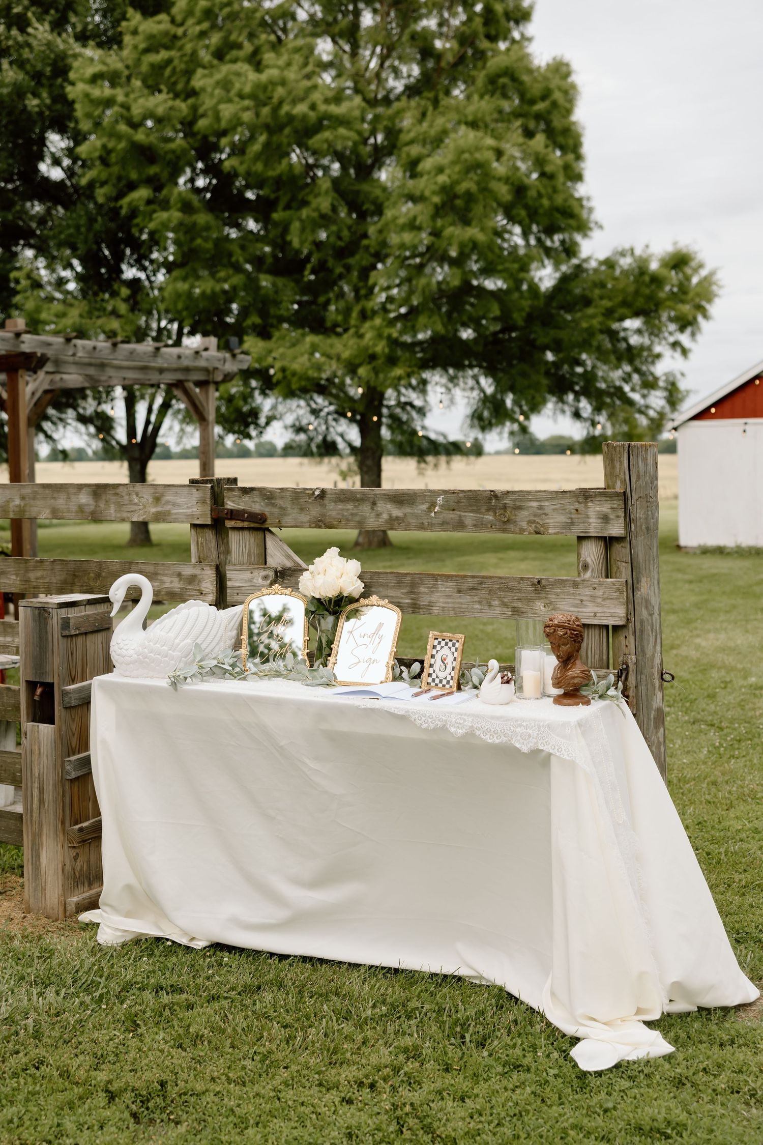 Elegant outdoor wedding gift table with white linen, swan decor, and framed photos set against rustic wooden fence.